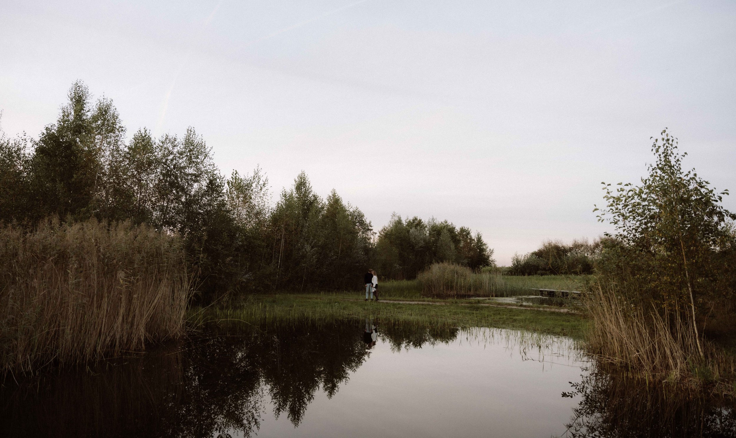 Twee mensen staan bij een klein meer in een natuurlijke omgeving, omgeven door bomen en riet, onder een heldere lucht.