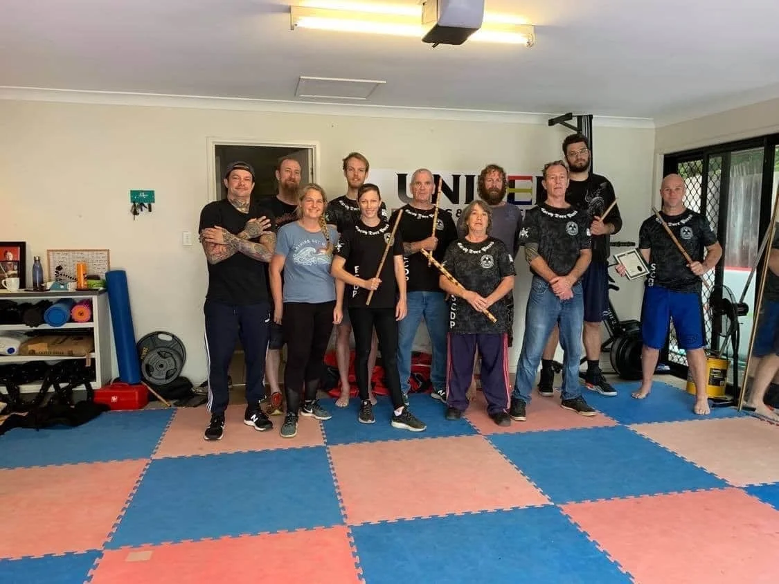 Group of people in martial arts uniforms and casual clothing posing in a martial arts studio with foam mats, fitness equipment, and a UNIM logo on the wall.