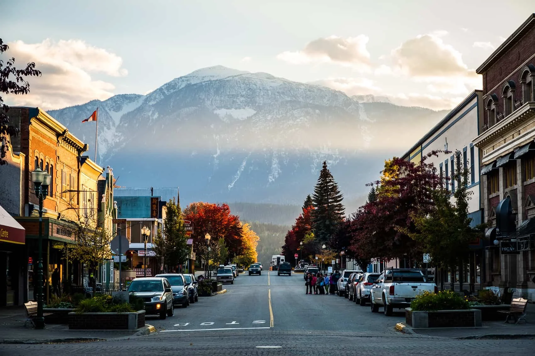 Street view of a small town with colorful buildings, parked cars, and trees with fall foliage, with mountains in the background and a partly cloudy sky.