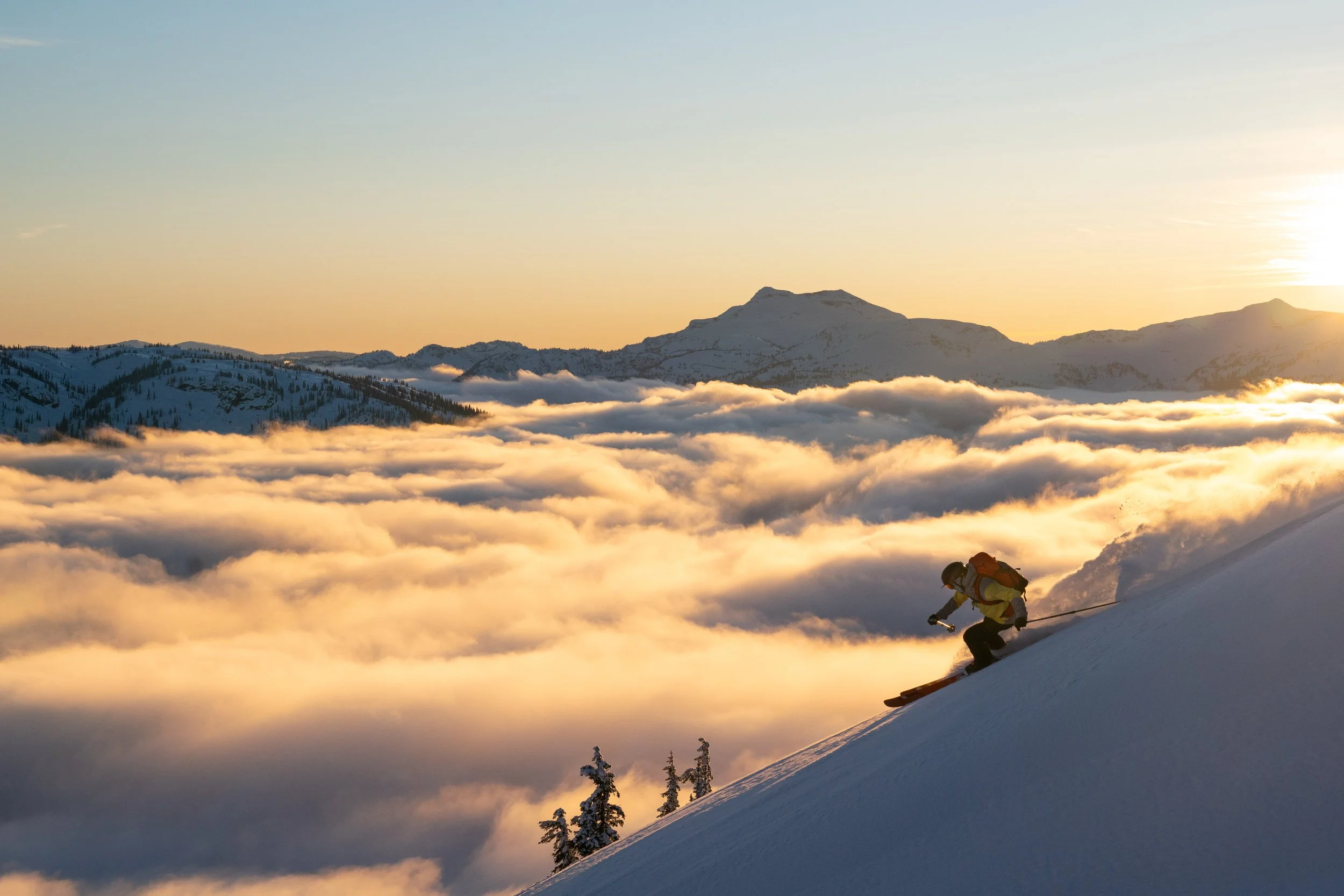 Skier descending a snowy mountain slope at sunrise above the clouds, with a picturesque mountain range in the background.