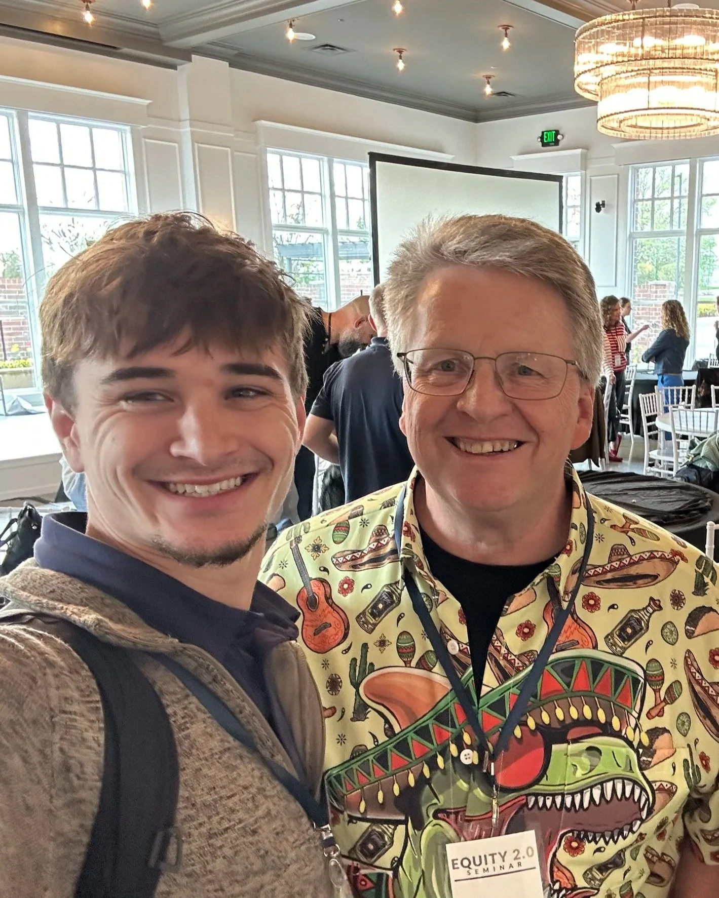 Two men smiling for a selfie indoors at a seminar or conference with large windows and a chandelier in the background. One man wears a gray hoodie and backpack, the other wears glasses and a colorful shirt with a dragon design and a seminar badge.