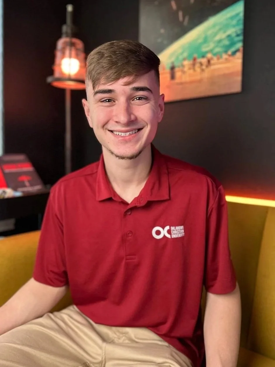 Young man with short brown hair and a goatee smiling, sitting on a yellow couch, wearing a red polo shirt with a logo, in a room with dark walls, a standing lamp, and artwork of Earth from space in the background.
