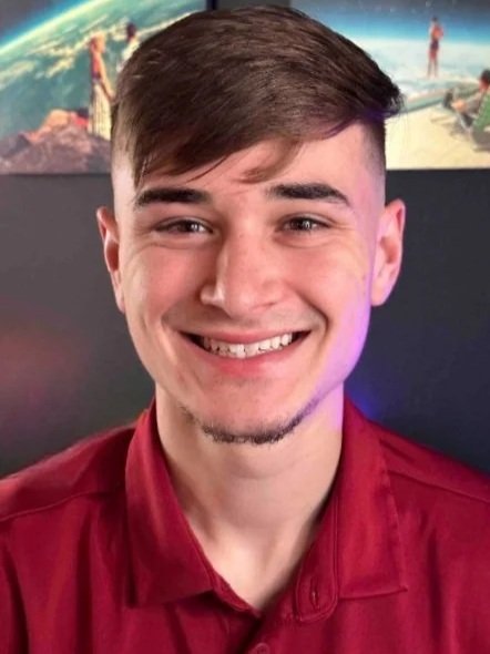 A young man with light brown hair and a short beard, smiling, wearing a red shirt, with a world map or globe in the background.