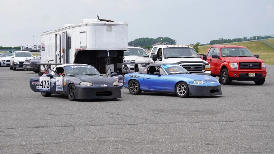 Two race cars parked on a race track with support vehicles and trailers nearby.