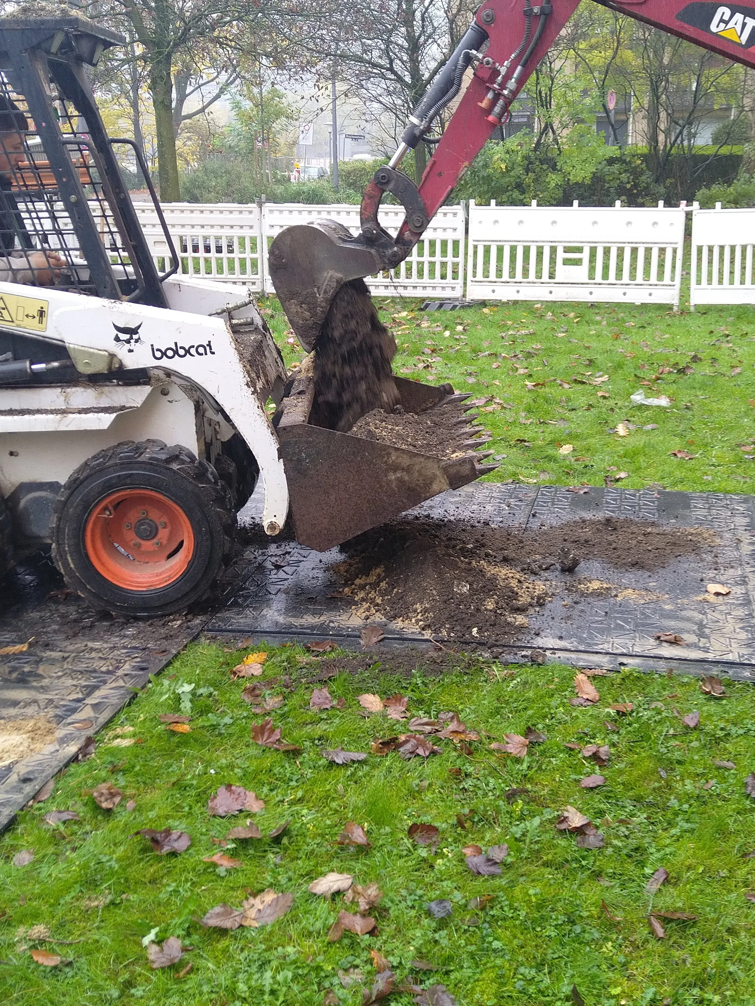Ein Bagger hebt Erde auf, während er auf Rasen mit Herbstlaub arbeitet. Im Hintergrund eine weiße Zäunung und Bäume.