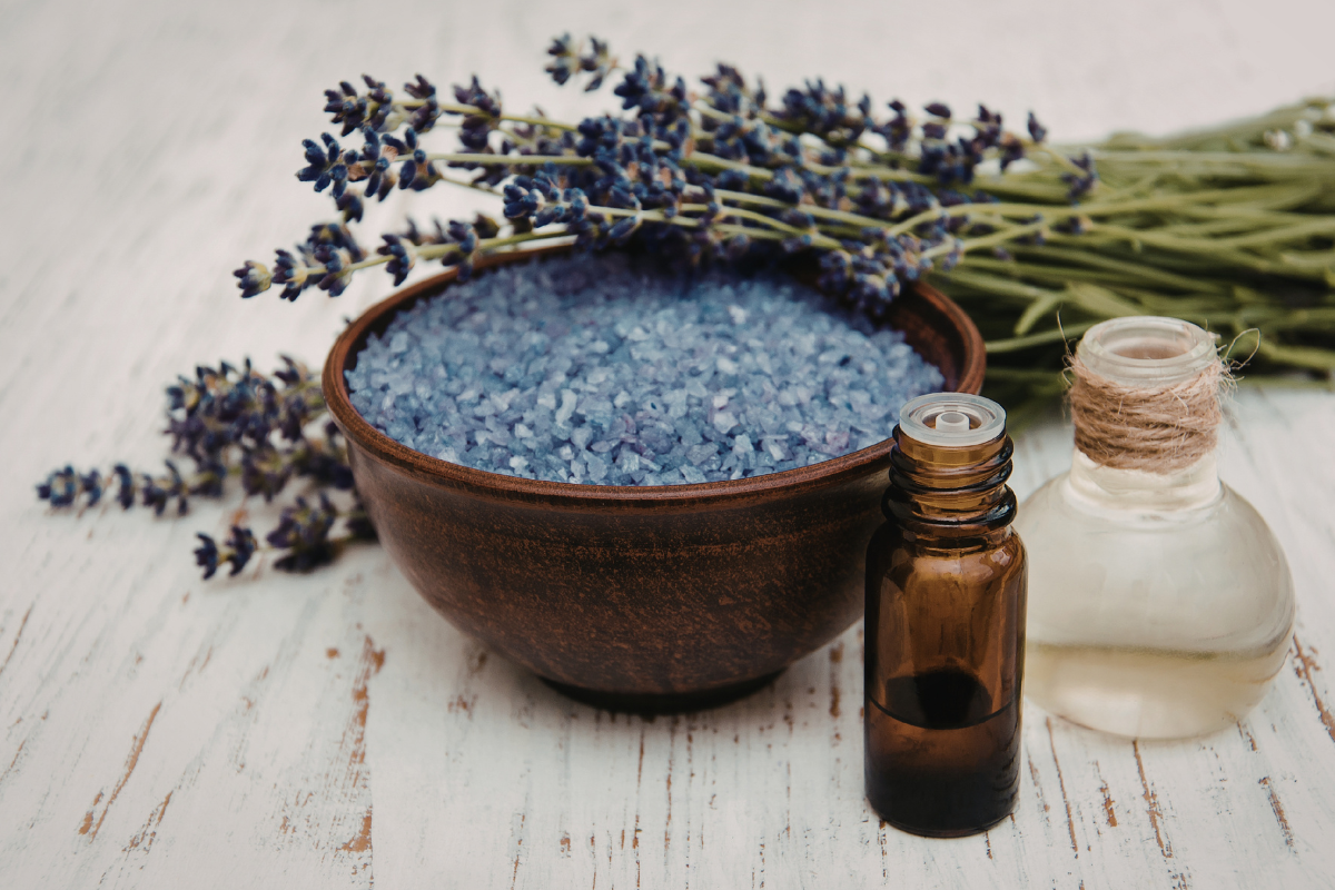 Zwei kleine Braunglasflaschen mit ätherischem Öl, eine Schale mit blauen Badesalzen und ein Bund frischer Lavendel auf weißem Holz.
