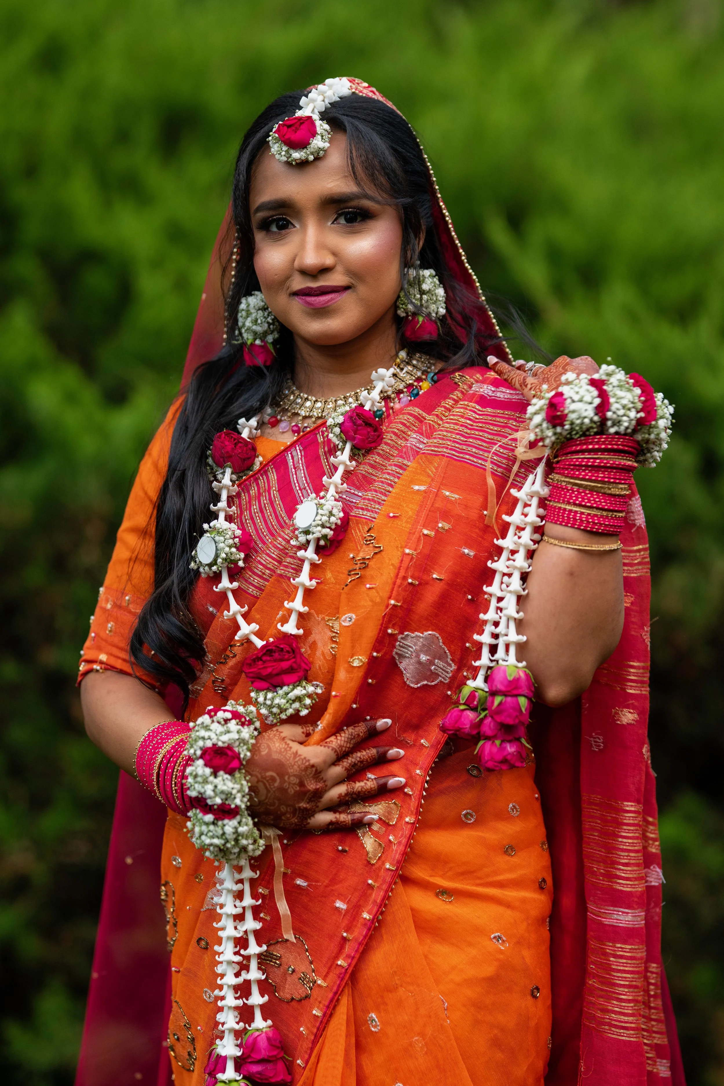 A woman dressed in traditional orange and pink saree, adorned with jewelry and flower garlands, standing outdoors against a green background.