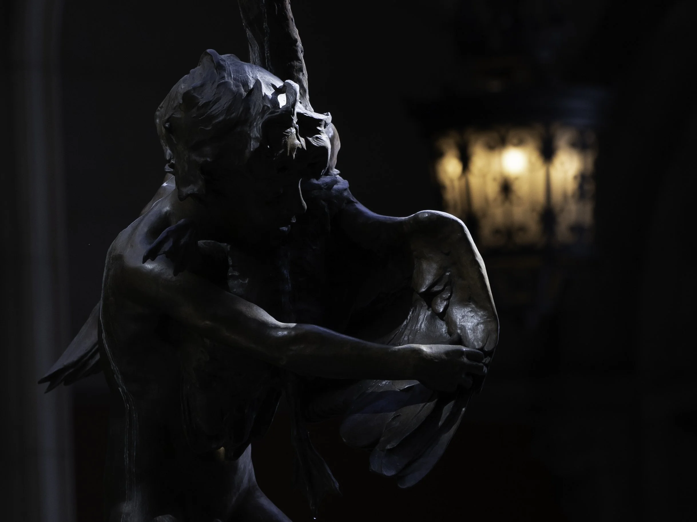A bronze statue of a man with long hair and beard, resting his chin on his hand, in a dark indoor setting with warm lighting in the background.