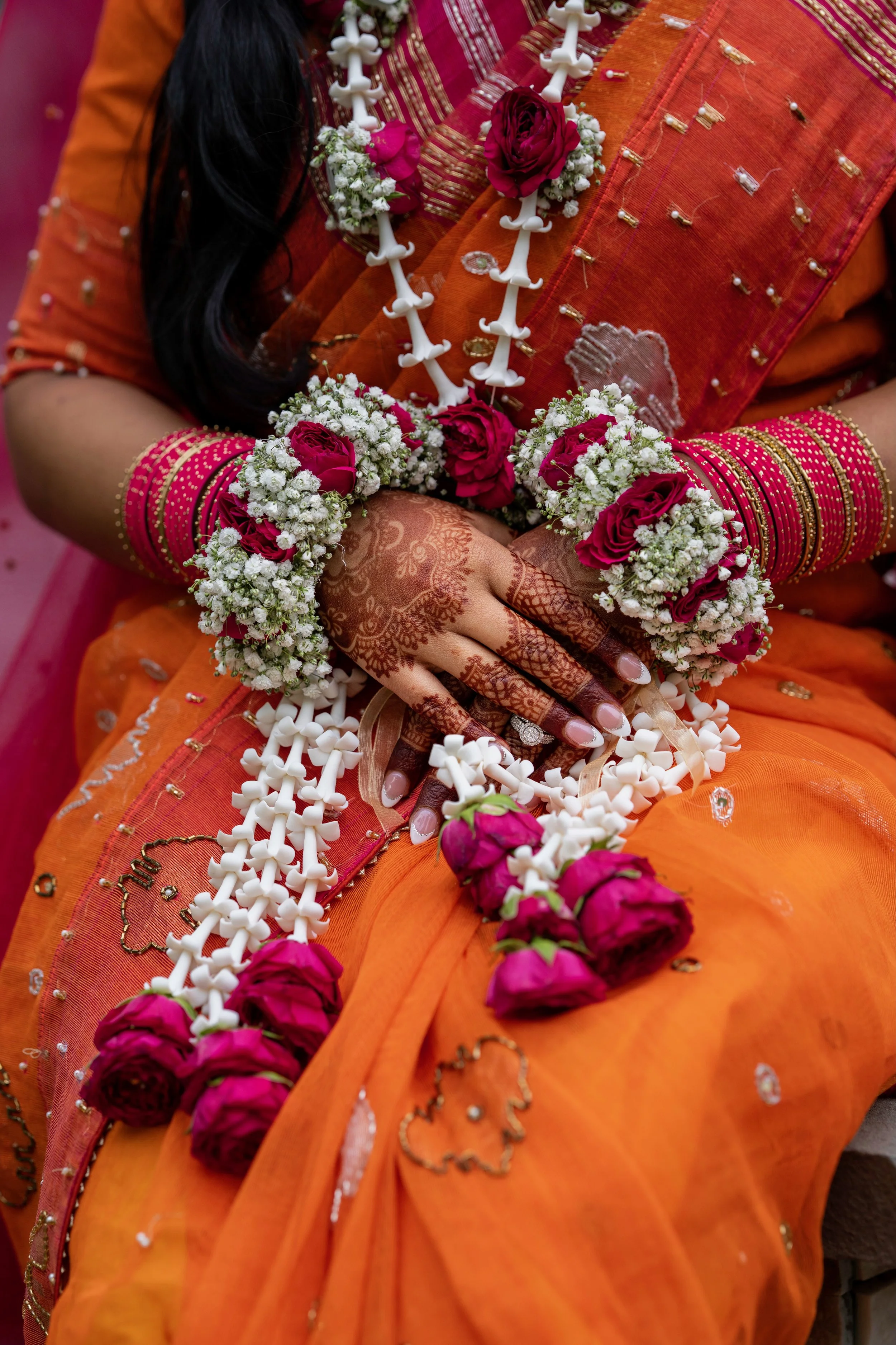 Close-up of an ornate orange traditional Indian sari decorated with beads and embroidery, adorned with pink roses, white flowers, and jewelry, with henna decorated hands resting on the lap.