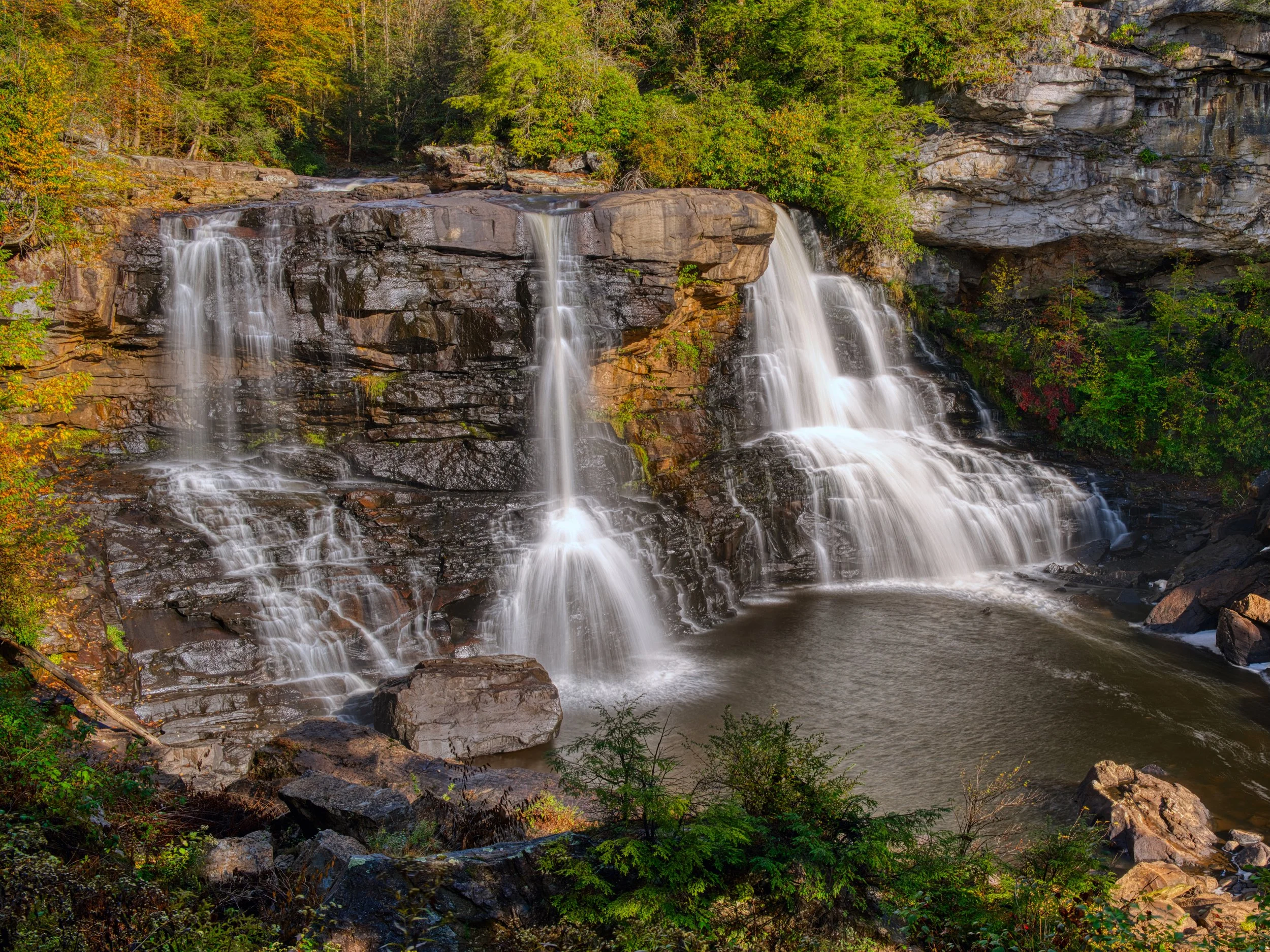 A multi-tiered waterfall cascading over rocks in a forest during autumn, with green, orange, and yellow foliage surrounding it.