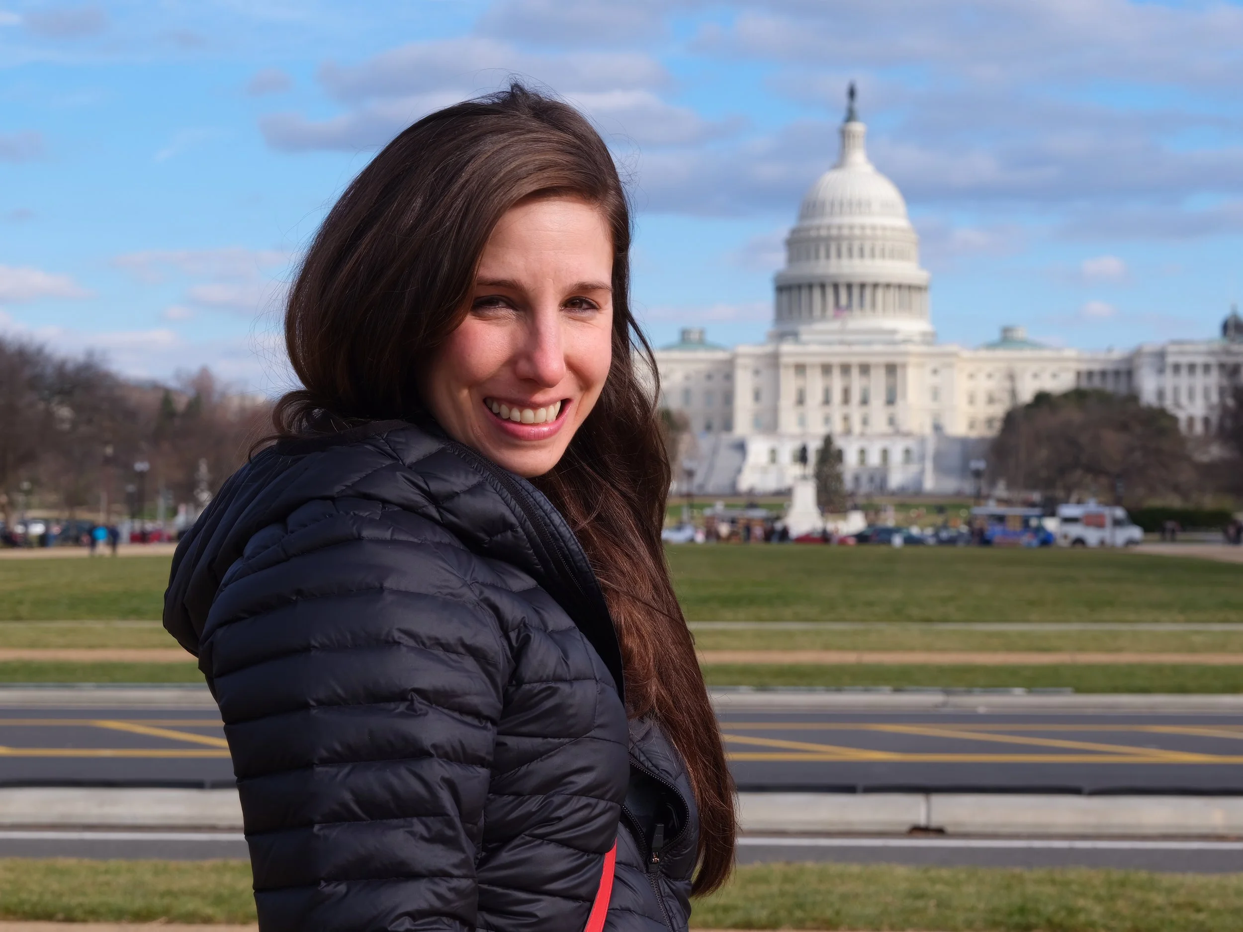 A young woman with long brown hair and a black puffer jacket smiling outdoors with the US Capitol building in the background on a partly cloudy day.