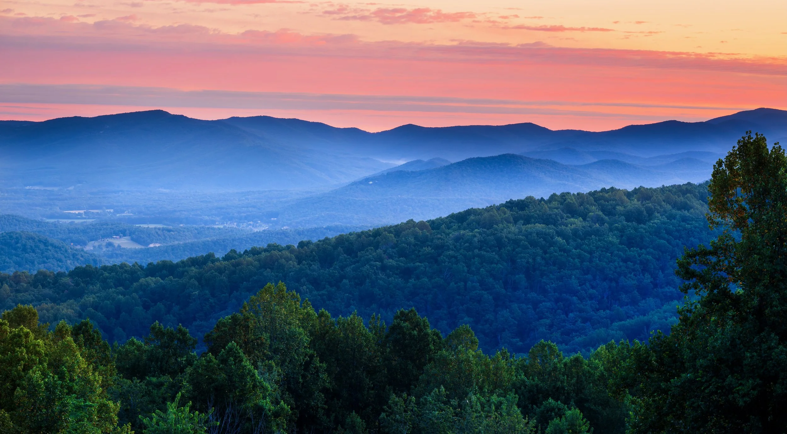 A scenic landscape of rolling mountains covered with dense green forests under a colorful sunset sky with pink and orange hues.