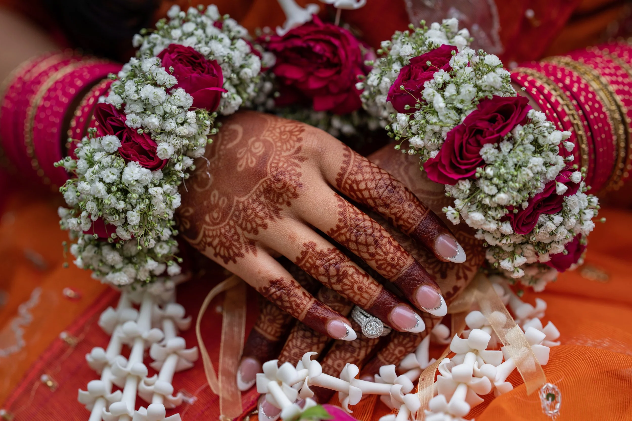 Close-up of a woman's hand with intricate henna designs, wearing rings, holding a flower garland of red roses and white baby's breath, surrounded by orange and beige fabric with decorative patterns and small white objects.