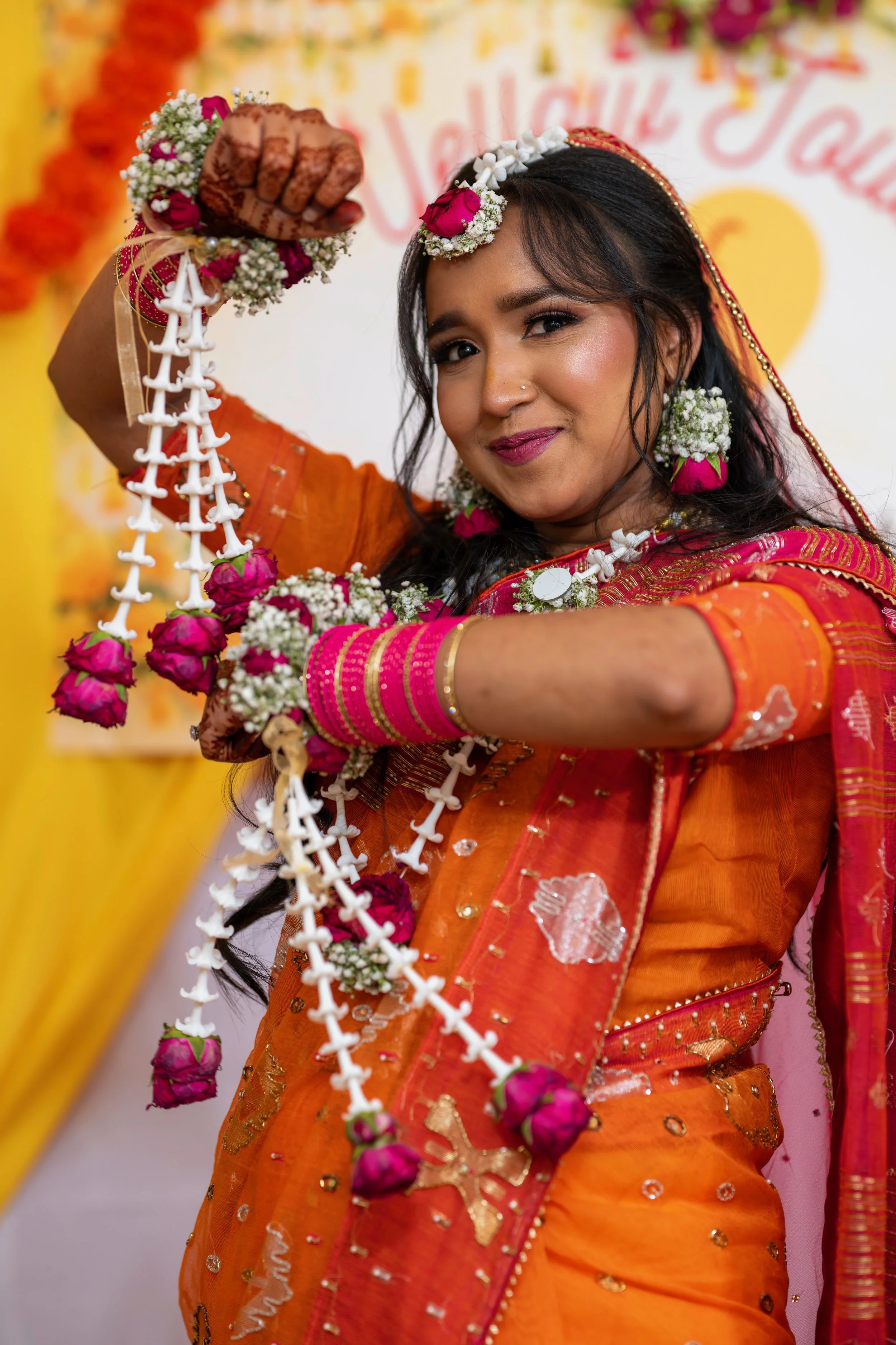 A woman dressed in colorful traditional Indian attire and jewelry, holding a flower garland, and smiling at the camera.