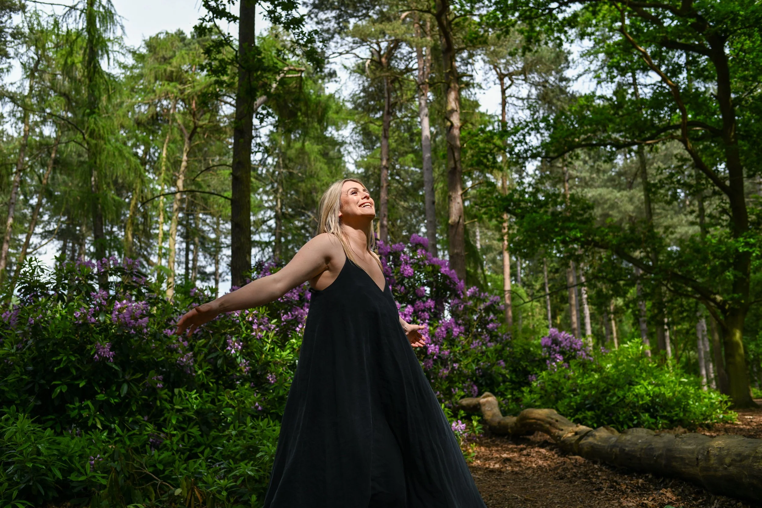 A woman in a black dress joyfully spreads her arms in a lush green forest with purple flowers and tall trees.