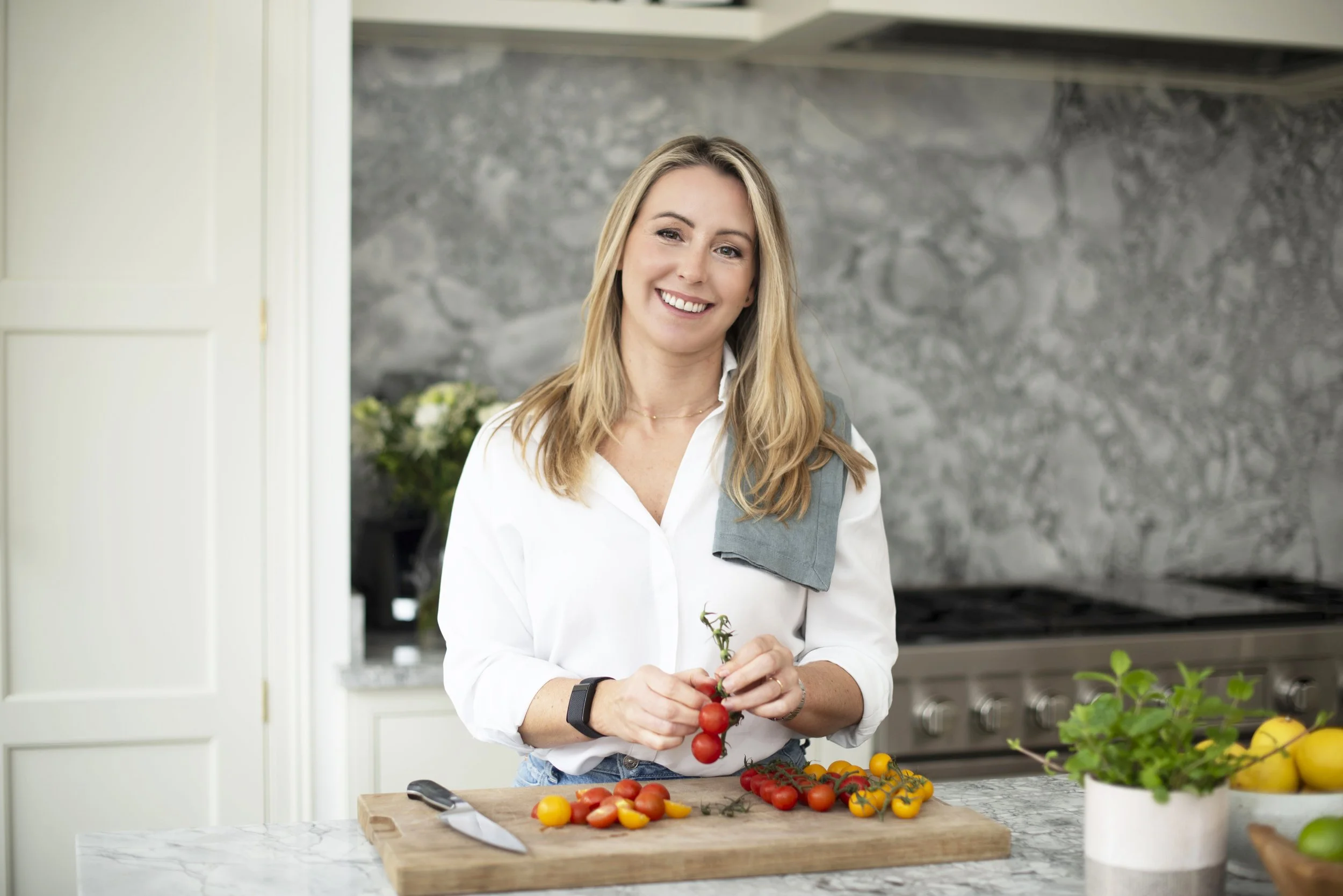 A woman smiling in a modern kitchen, preparing cherry tomatoes on a wooden cutting board.