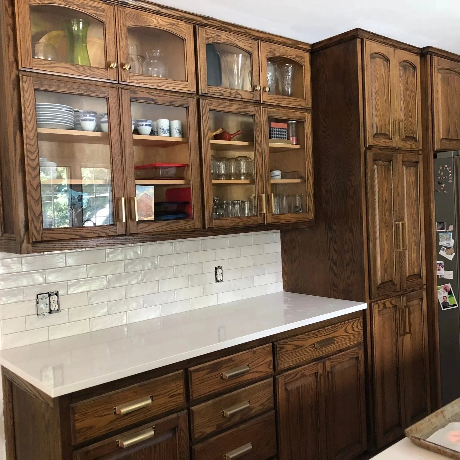 Kitchen cabinets with glass doors displaying dishes and glassware, white countertop, beige subway tile backsplash, partially visible refrigerator with photos and magnets.