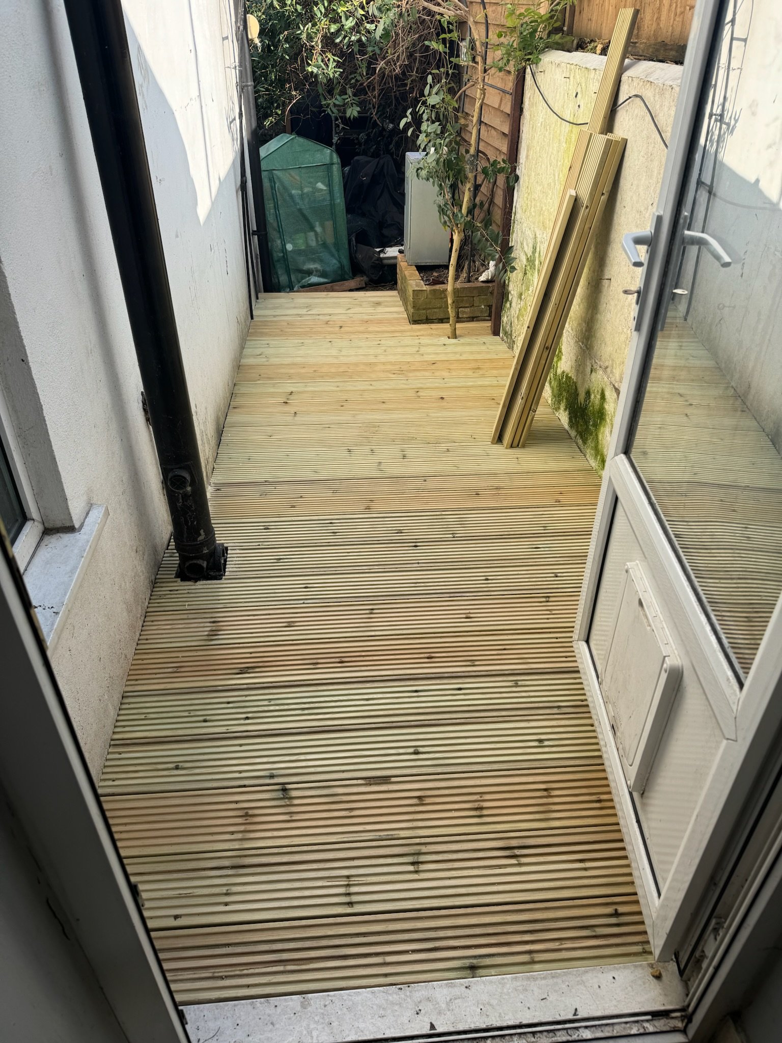 Newly installed wooden deck on a small outdoor patio, with construction materials stacked against a wall and plants in the corner.