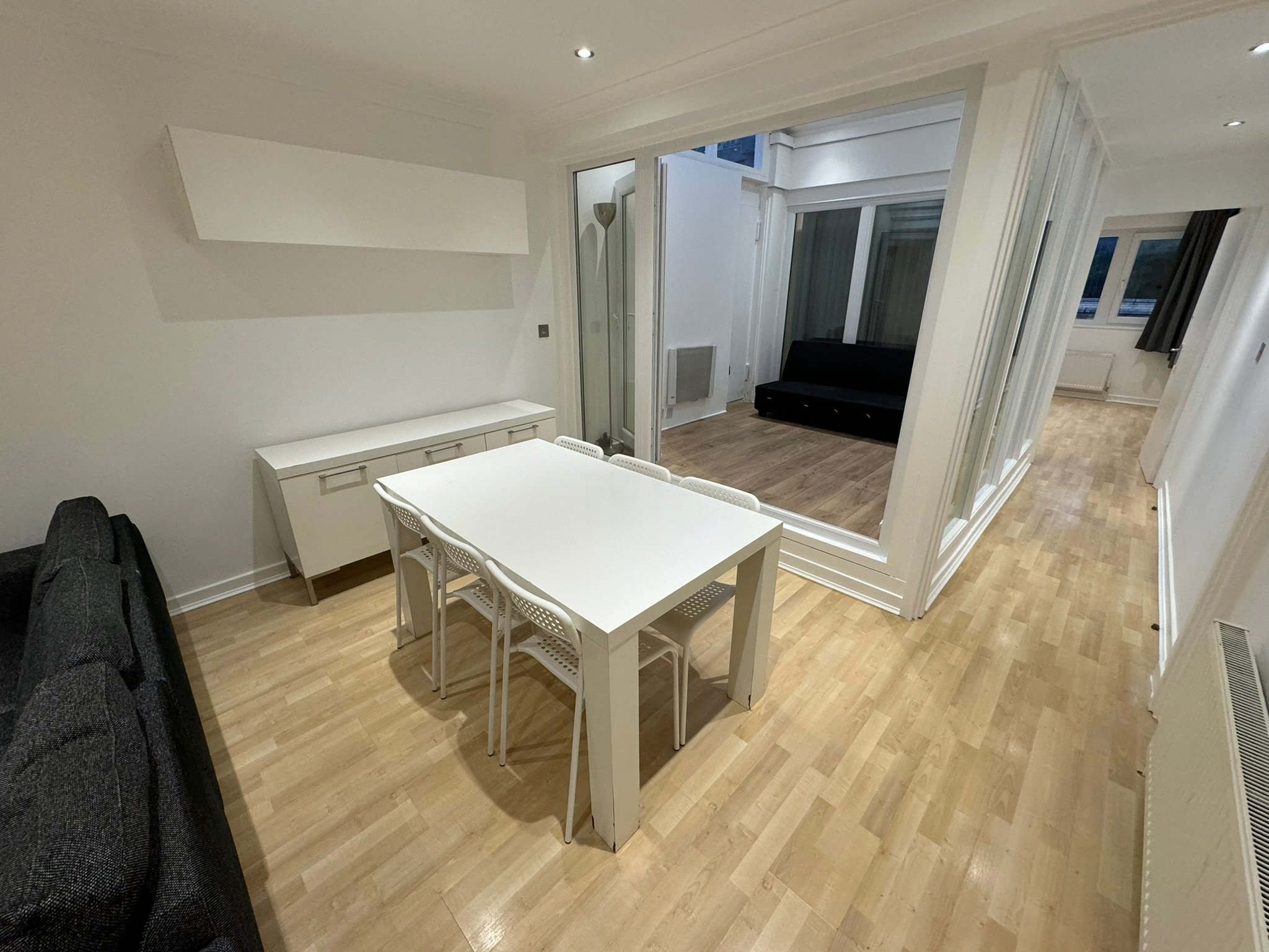 Dining area with white table and four white chairs, adjacent to a living space with dark sofa and large windows, in a modern apartment with light wood flooring.