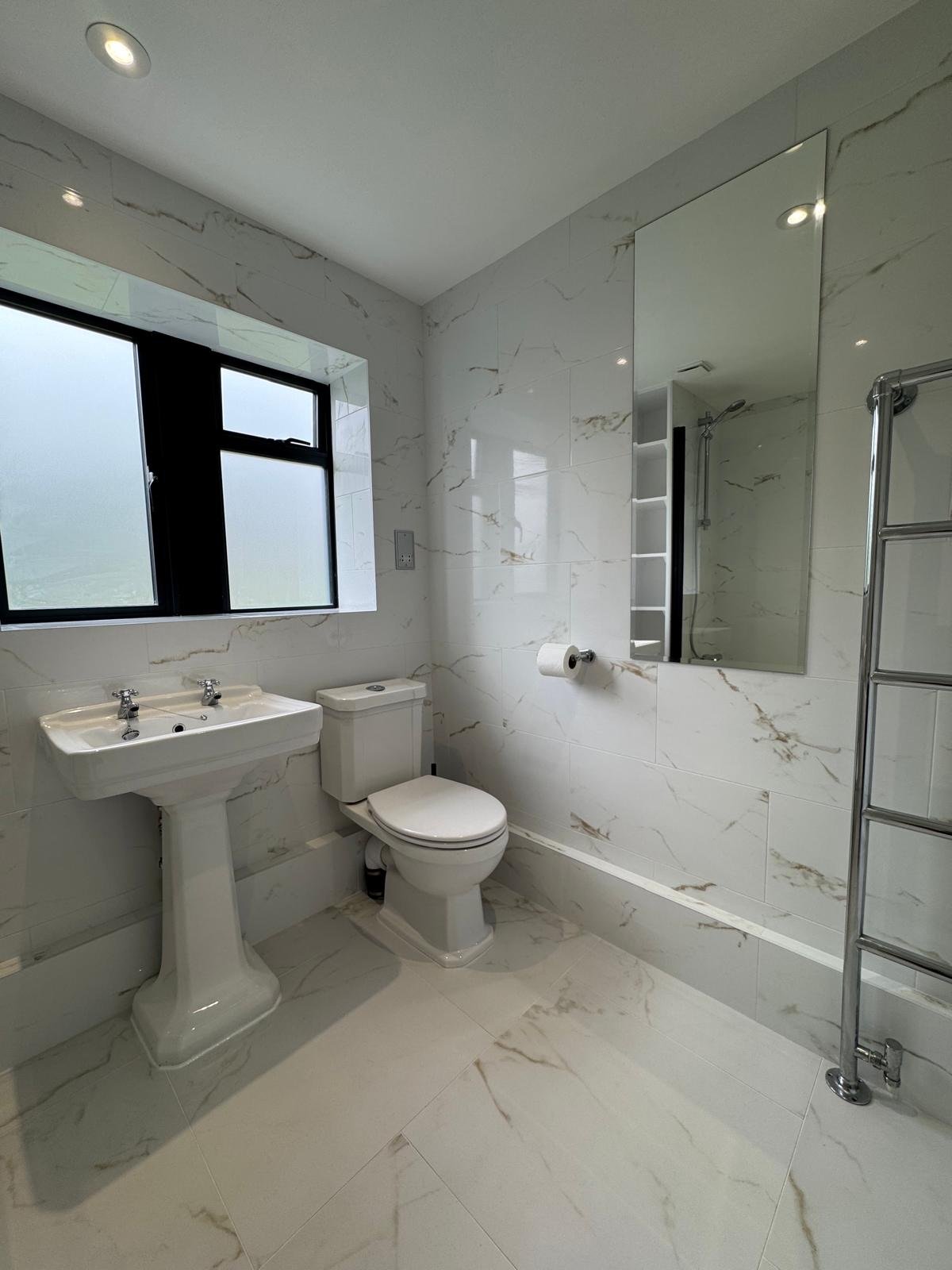 Bathroom with white marble walls and floor, black-framed window, a toilet, and a double sink pedestal, large mirror, and chrome towel rack.
