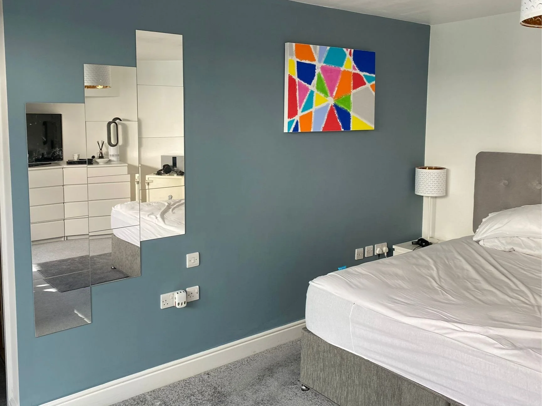 A bedroom with a bed, a blue accent wall, a white nightstand with a lamp, a colorful abstract painting, a mirror, and a reflection of a dresser and part of the bed in the mirror.