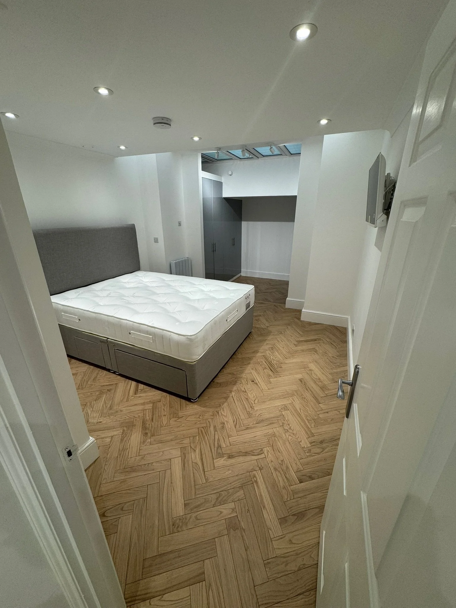 A bedroom with white walls and ceiling, featuring recessed lighting, a gray upholstered bed with a mattress, wooden herringbone-patterned flooring, a small white radiator, gray built-in cupboards and a skylight.
