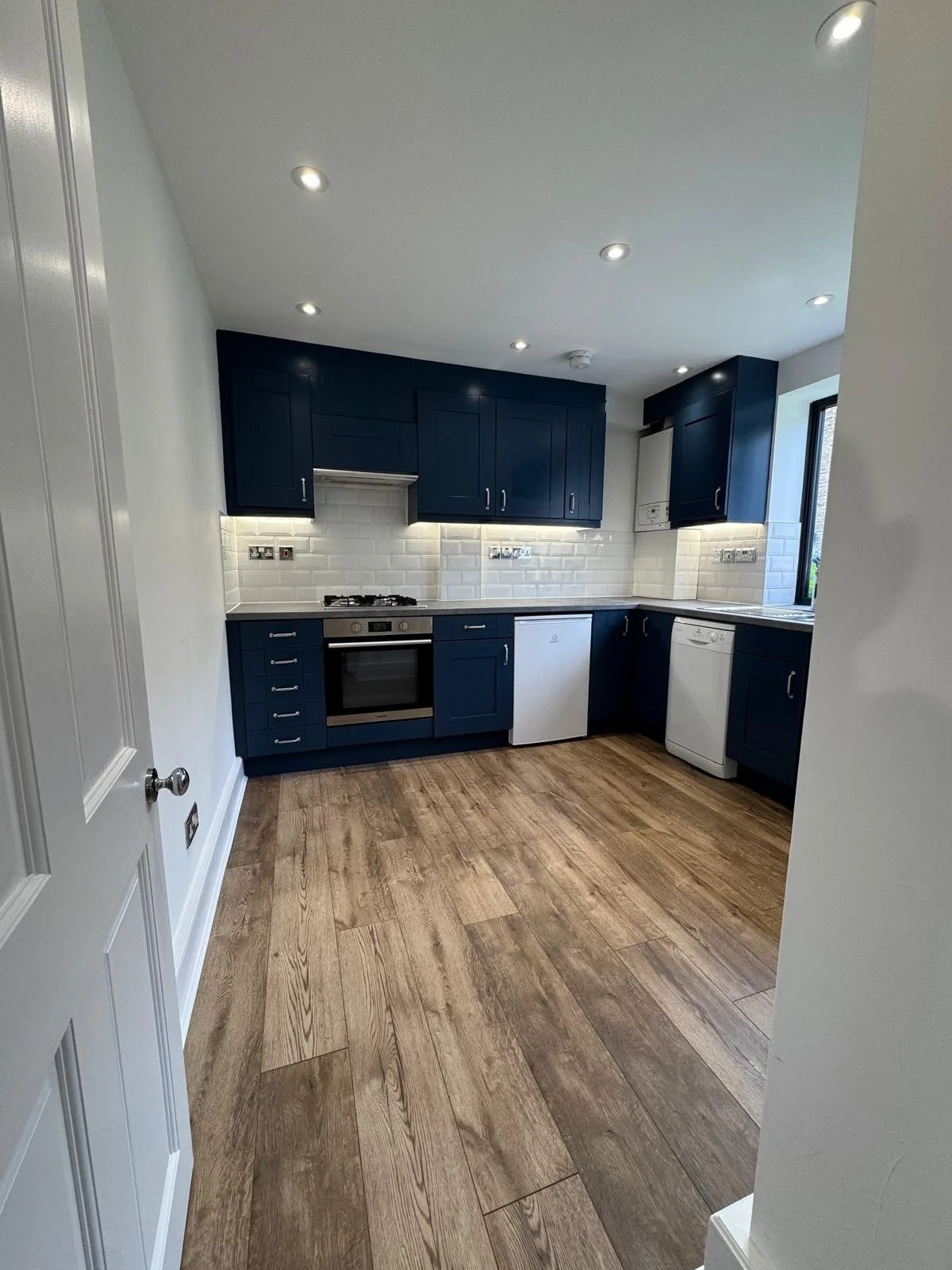 Modern kitchen with white subway tile backsplash, navy blue cabinets, stainless steel oven, white dishwasher, small refrigerator, and natural wood flooring.
