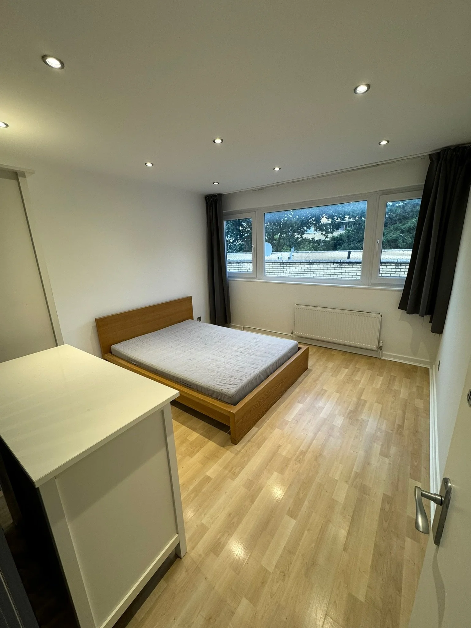 Empty bedroom with wooden bed frame and mattress, white dresser, large window with dark curtains, ceiling lights, hardwood floor, radiator under window.