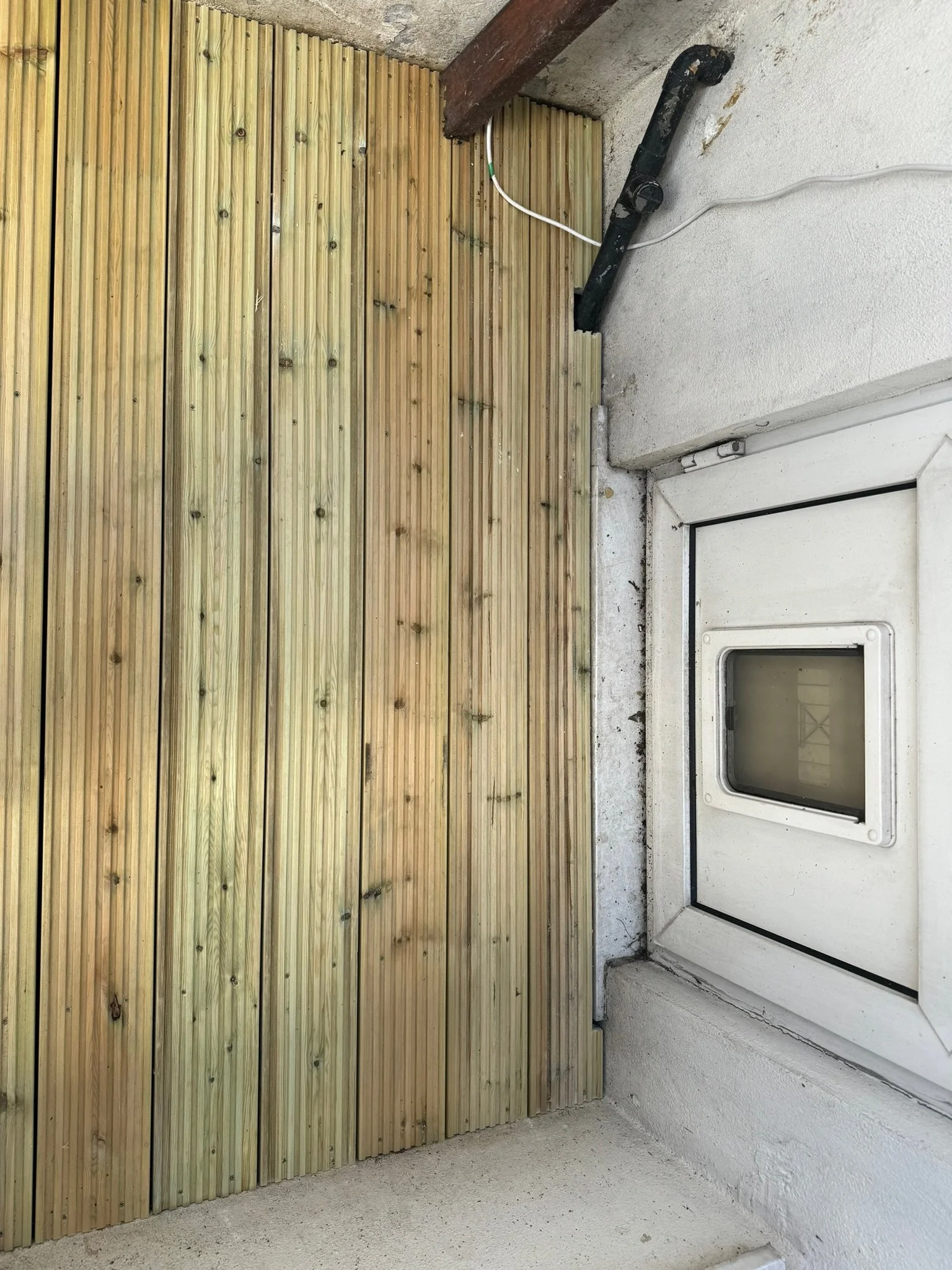 A corner of a room with a white ceiling and concrete wall. A wooden slat wall on the left side. A small white window or hatch on the right side with a black border. There is a black metal pipe and some cables near the ceiling.