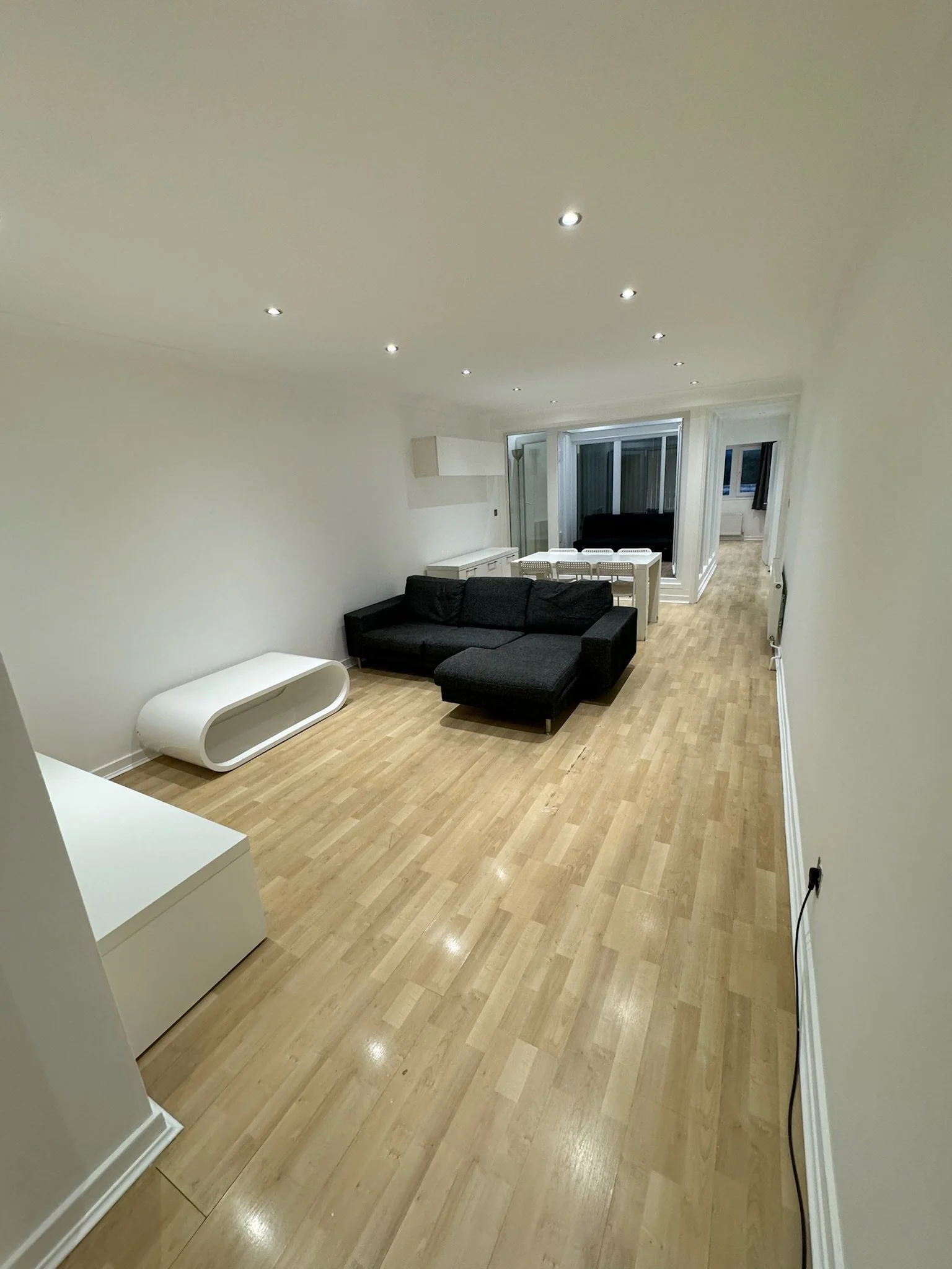 Empty living room with black sectional sofa, white coffee table, light wooden floor, and white walls, illuminated by ceiling spotlights.