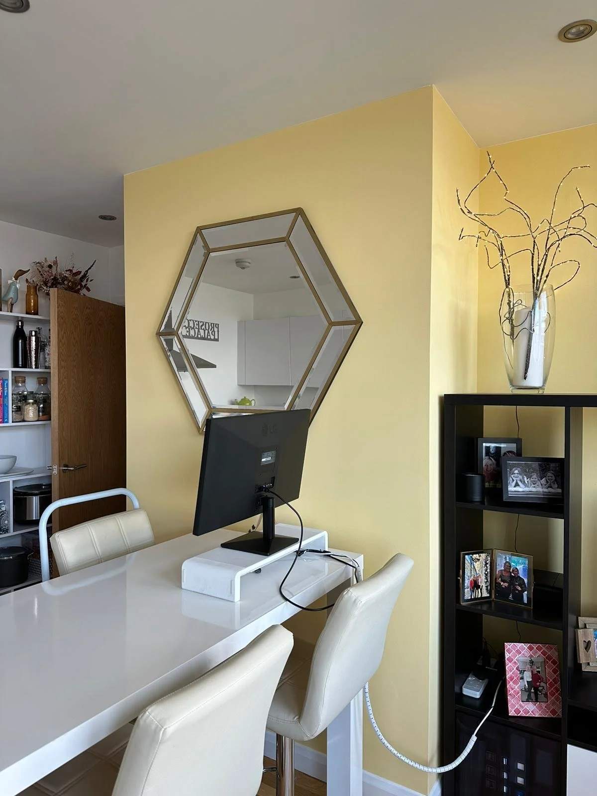Interior of a dining area with a white dining table, cream-colored chairs, a monitor, a hexagonal mirror on a yellow wall, and a black shelf with framed photos, a vase with decorative branches, and other items.