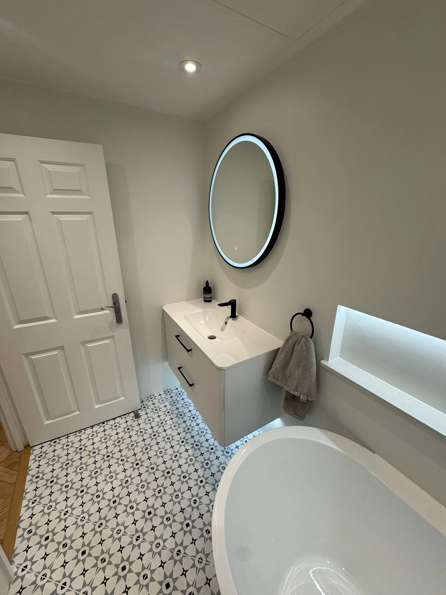 Modern bathroom with a round backlit mirror, a white vanity sink, a gray towel on a ring, a black faucet, patterned floor tiles, and a bathtub.