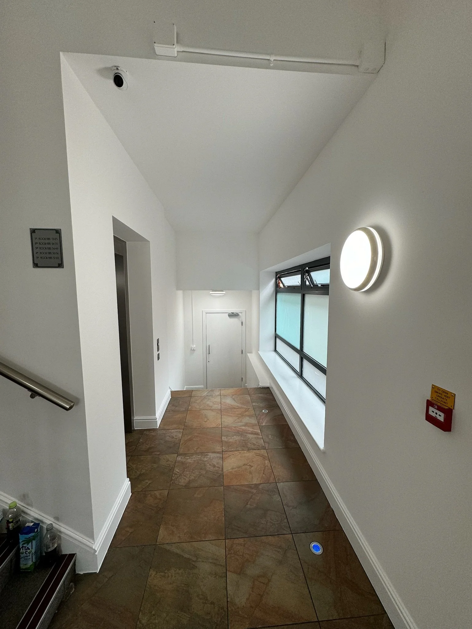 Interior view of a stairwell landing with a tiled floor, white walls, a window, and an elevator door on the left. There is a wall-mounted round light fixture on the right.