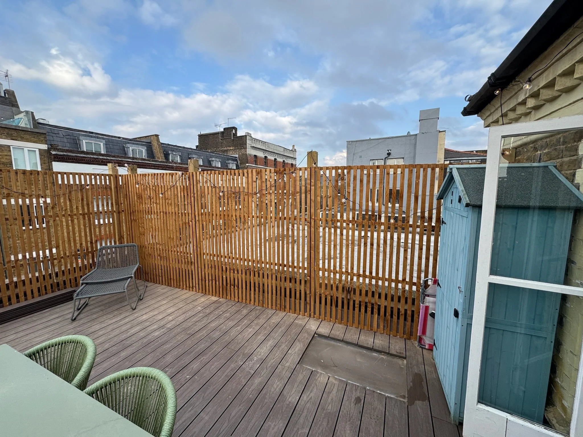 Urban rooftop terrace with wooden decking, a gray metal chair, and a blue shed, surrounded by a tall wooden fence under a partly cloudy sky.