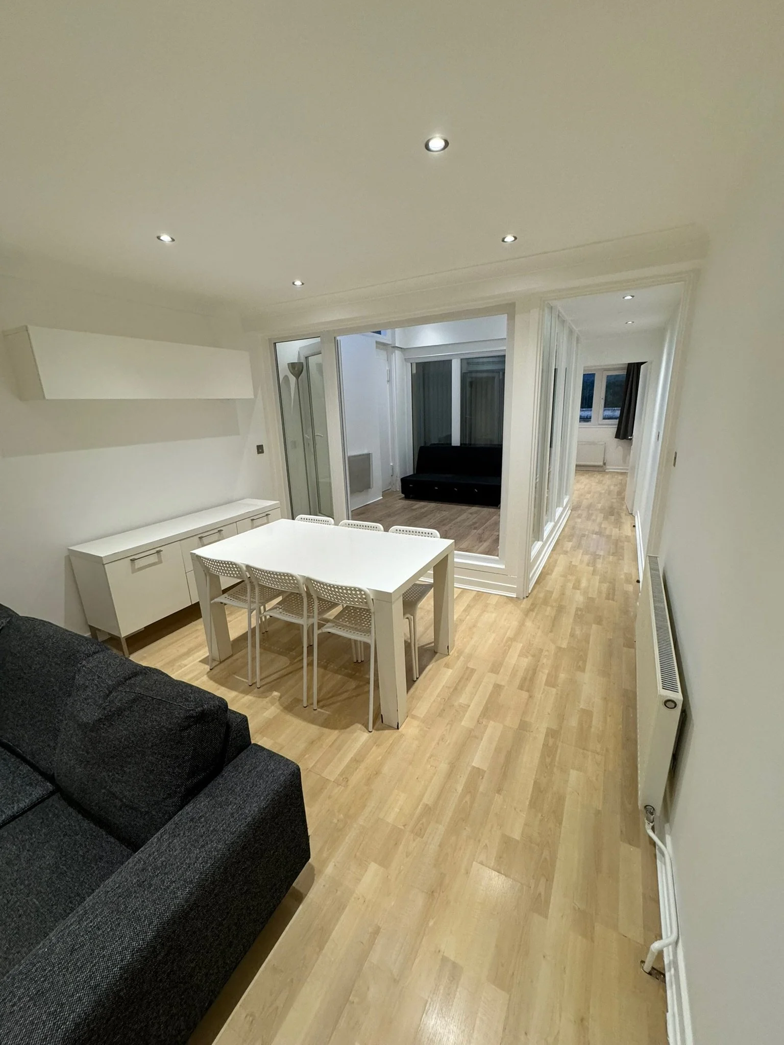 Living room with black sofa, white dining table with four white chairs, wooden flooring, and a small sitting area with a black couch visible through glass doors.