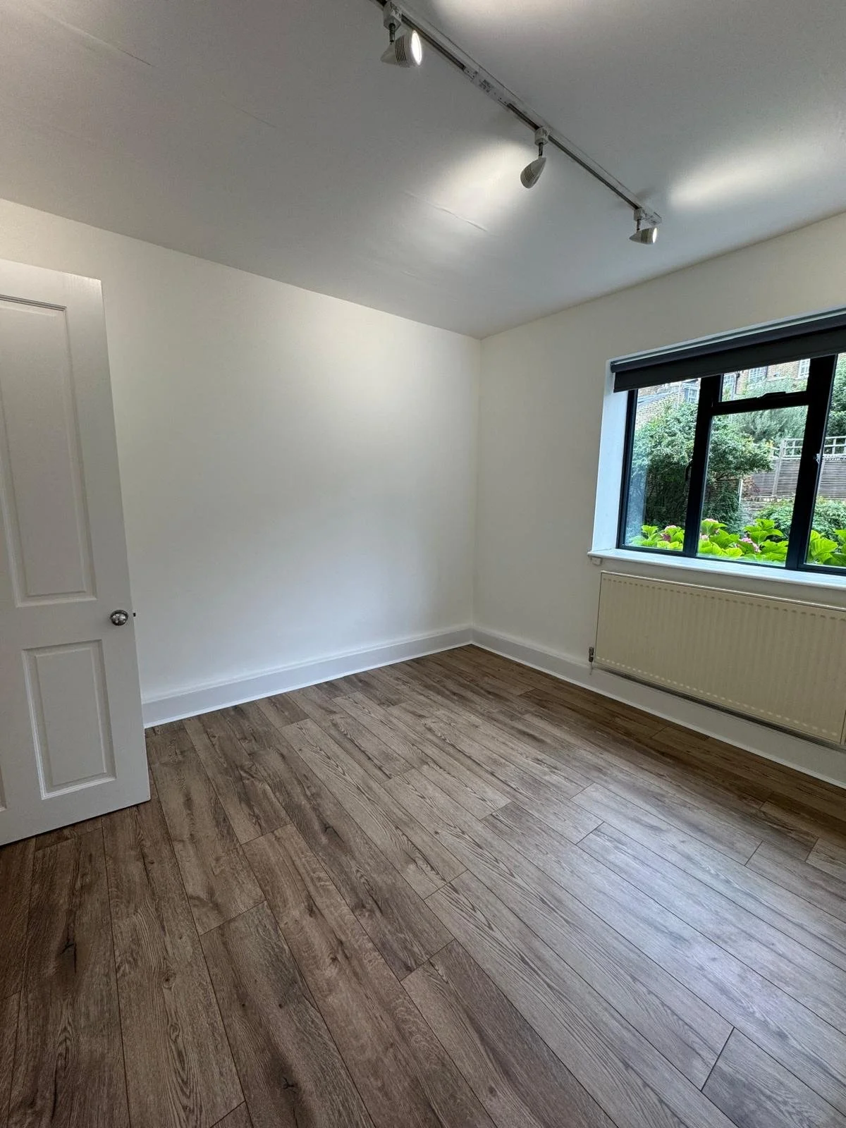 Empty bedroom with white walls, a window with a view of greenery, brown wood flooring, a white door, and ceiling spotlights.
