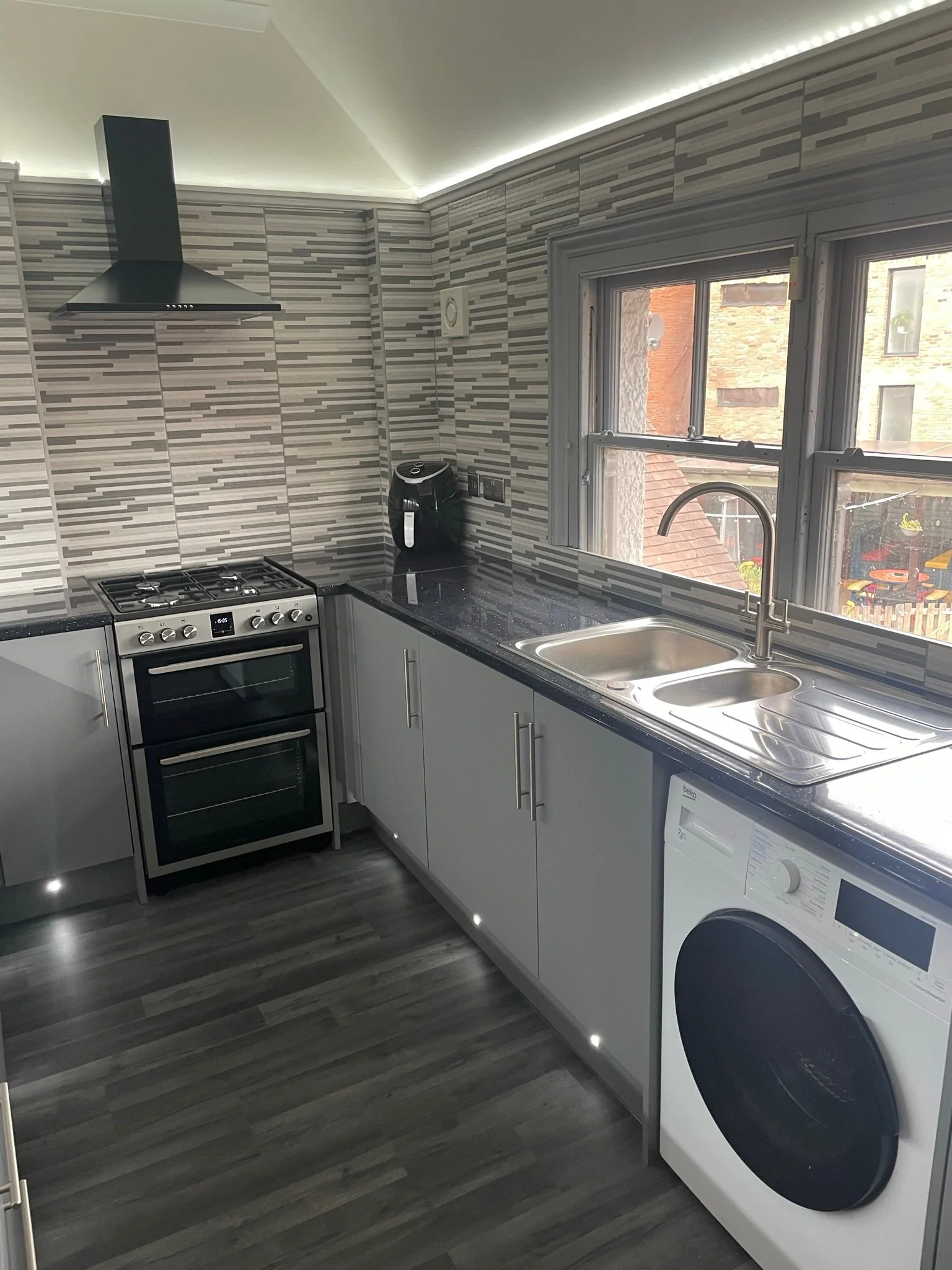Modern kitchen with gray cabinets, black countertop, stove, microwave, and a washing machine under the counter. There is a double sink next to a window, and a black coffee maker on the counter. The backsplash is striped gray and white tiles, with gra