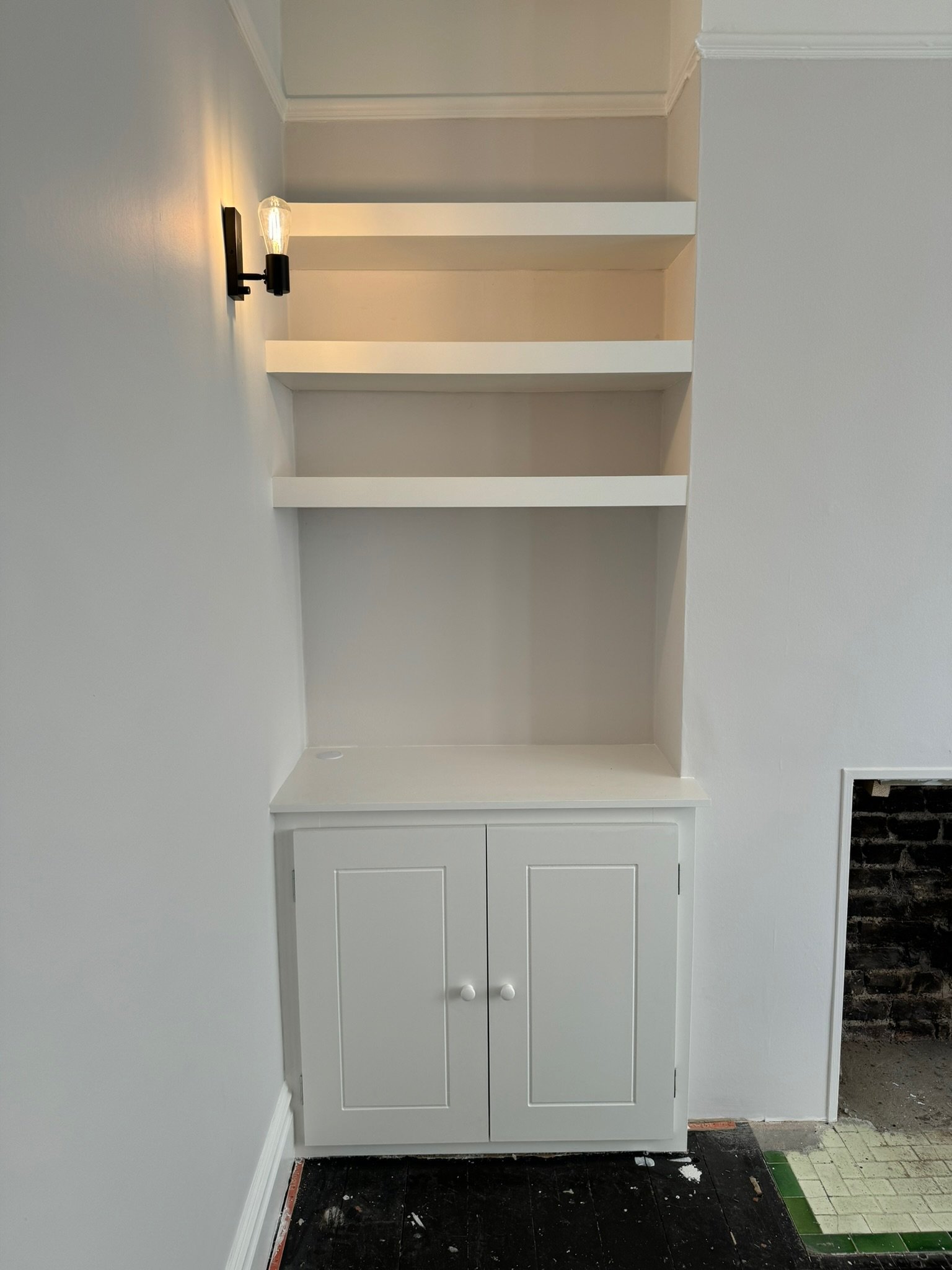 Built-in white cabinet with three open shelves and closed storage at the bottom, mounted on a white wall with a black wall sconce light to the left.