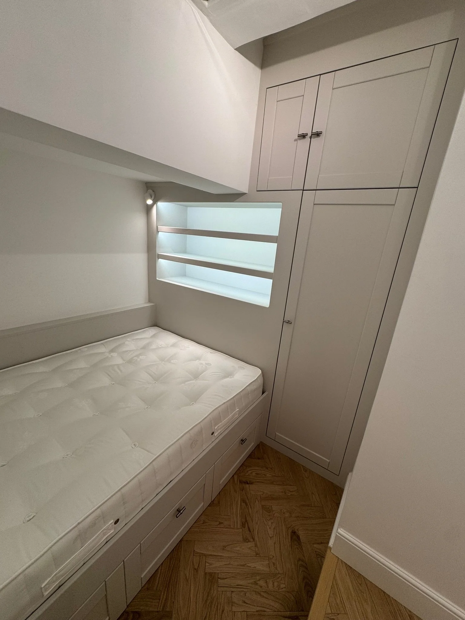 Small bedroom with white cushioned bed, built-in white wardrobe, and a window with three built-in shelves underneath. Wooden herringbone-patterned floor.