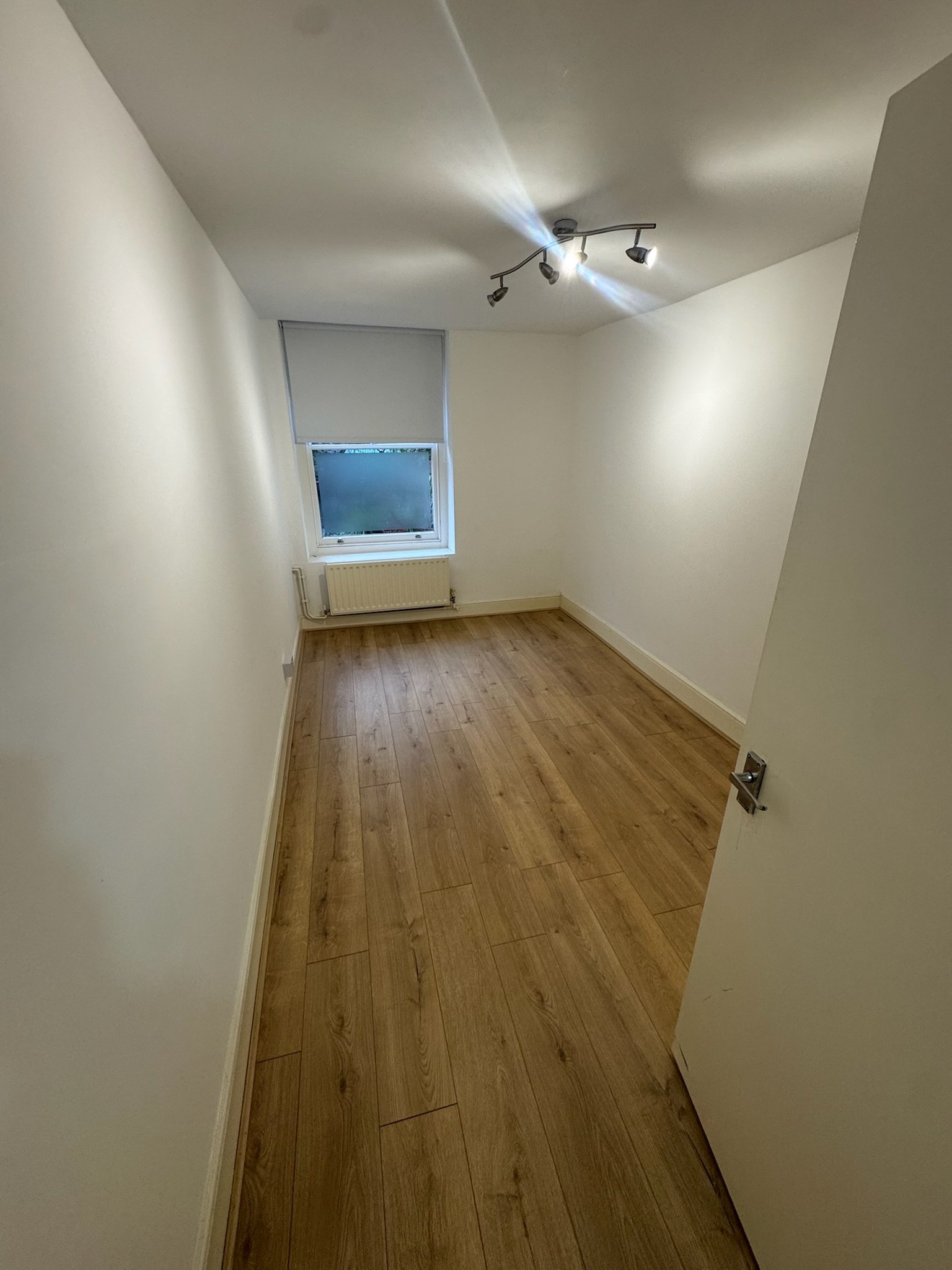 Empty small room with white walls, wooden floor, window with blinds, radiator, and ceiling light fixture.