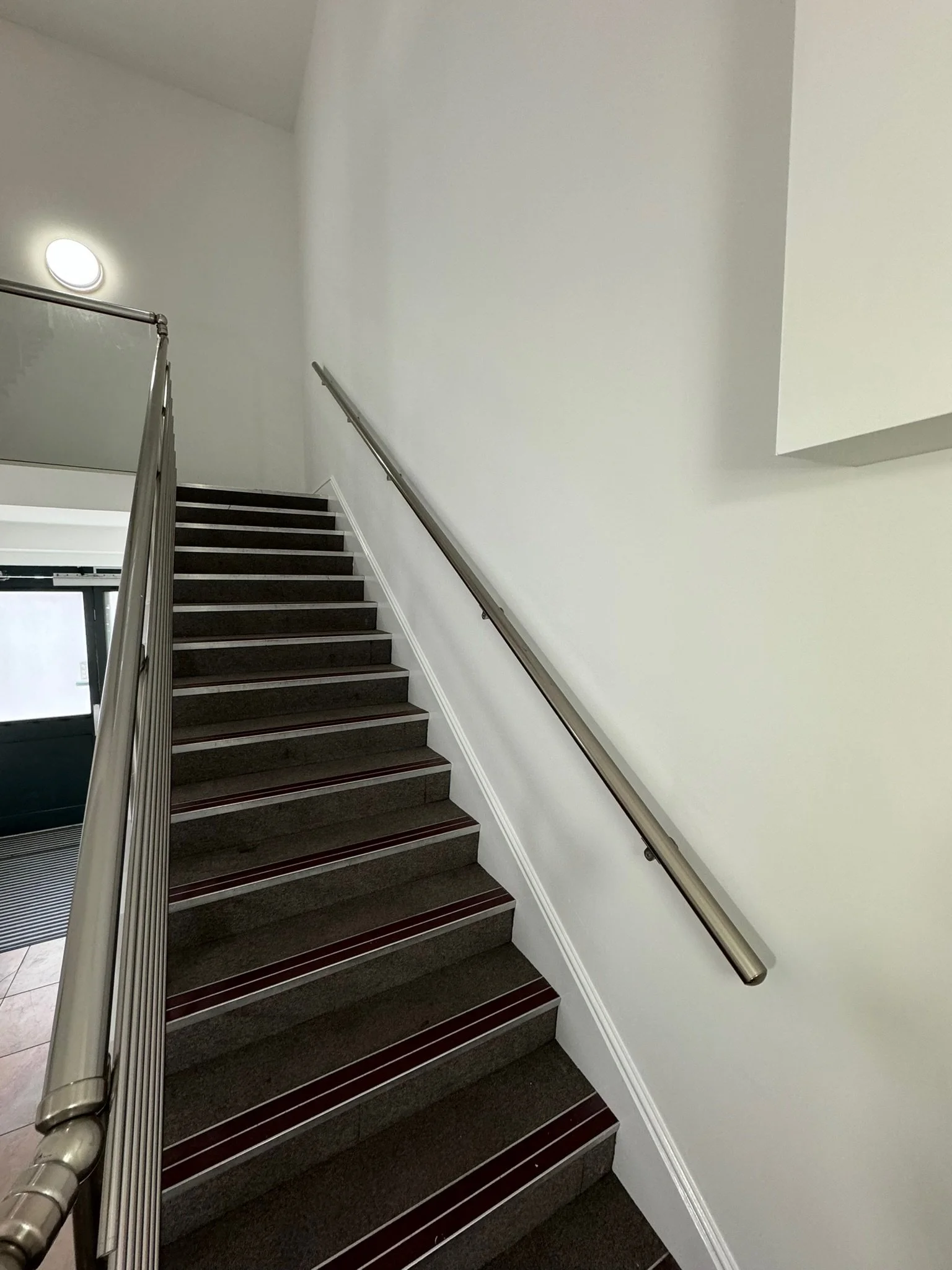 Staircase with black steps and a silver handrail in a modern interior with white walls and beige tile flooring.