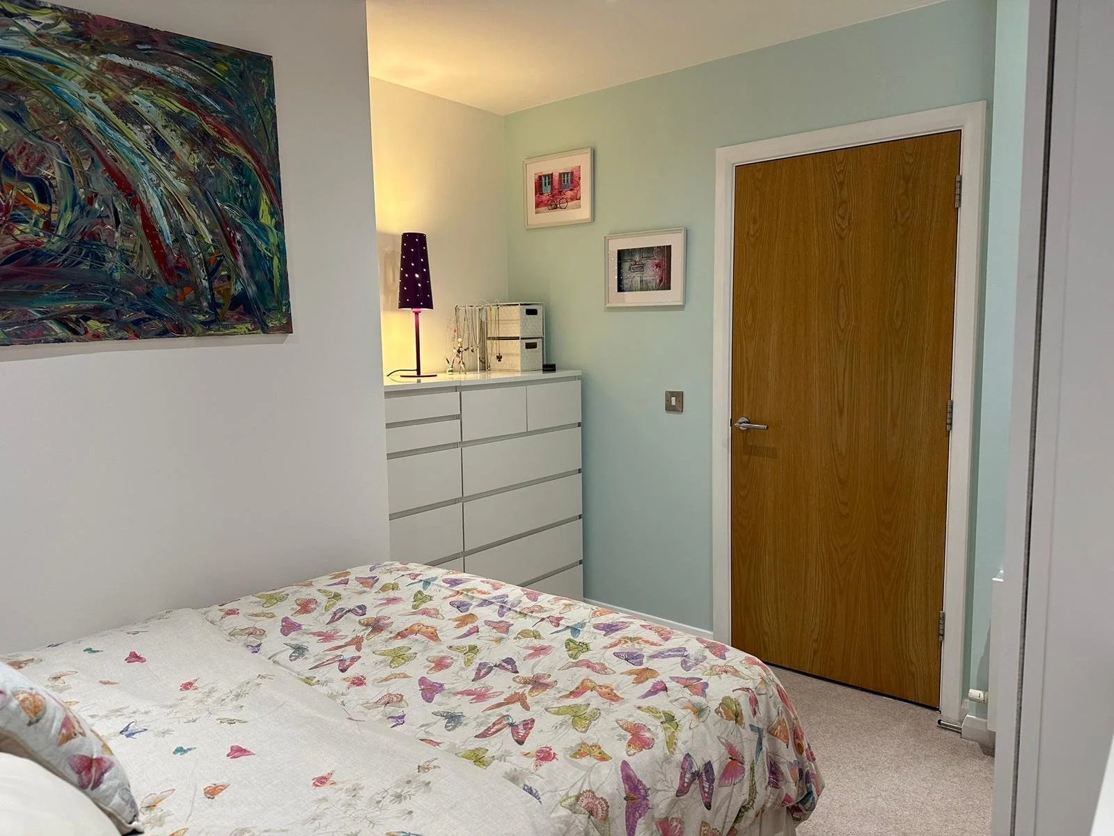 A bedroom with light green walls, a white dresser, colorful abstract art on the wall, and a bed with a butterfly-patterned bedspread.