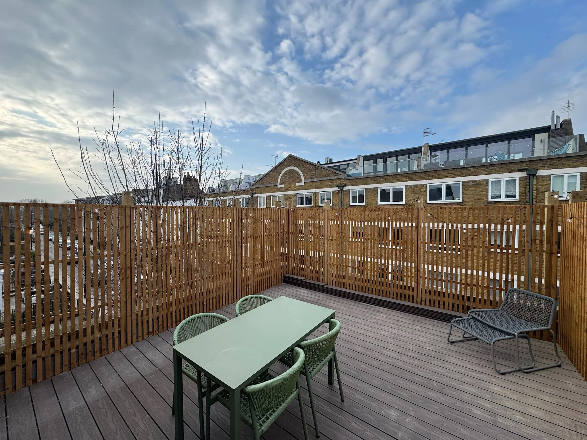 Rooftop terrace with wooden floor, a small table with four green chairs, and a lounge chair, surrounded by a wooden fence, with apartment buildings and a partly cloudy sky in the background.