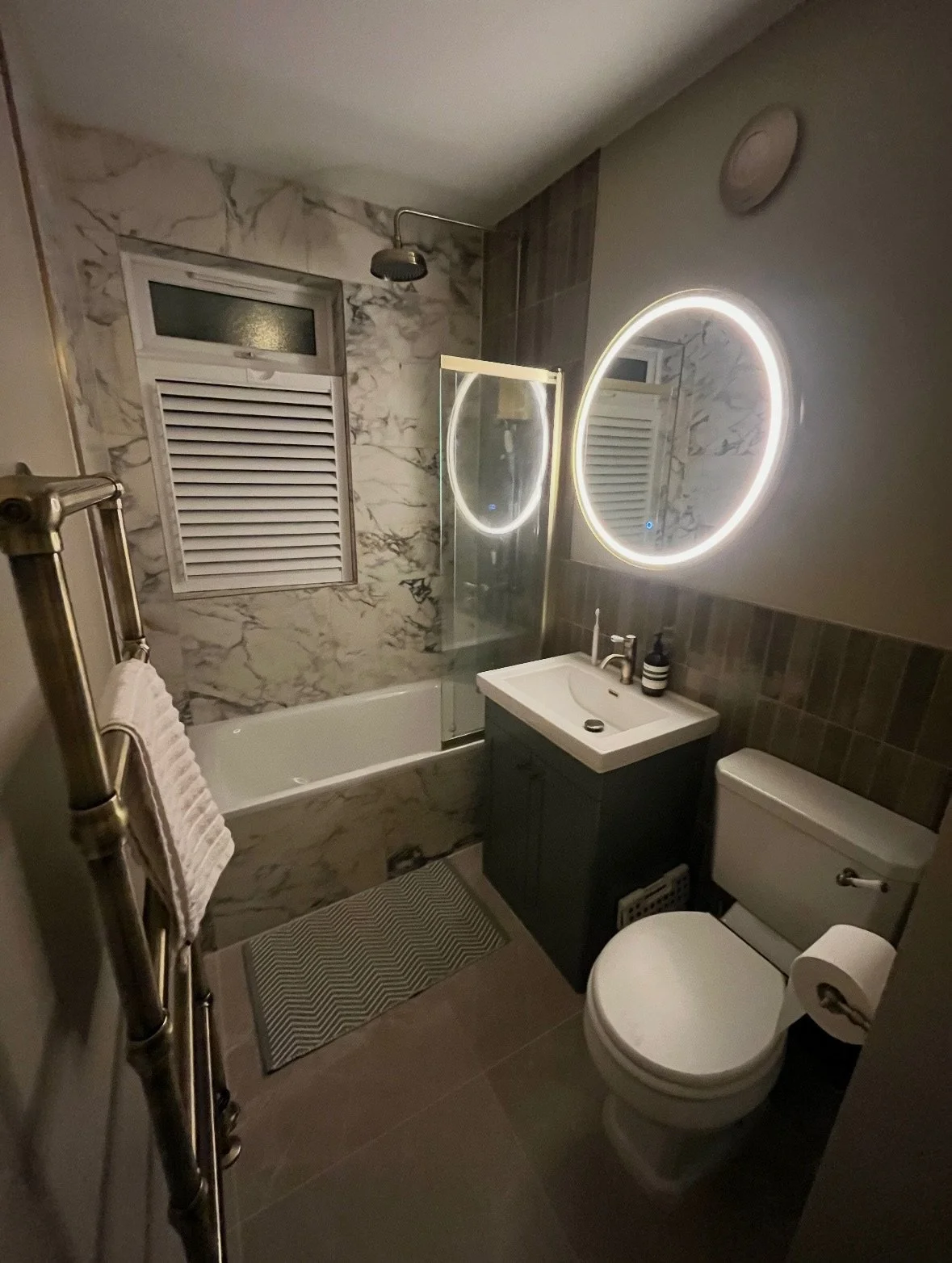 Modern bathroom with marble wall tiles, a bathtub with a glass divider, a wall-mounted round LED mirror, a small sink with a cabinet, a toilet, a window with a vent, and a towel rack.