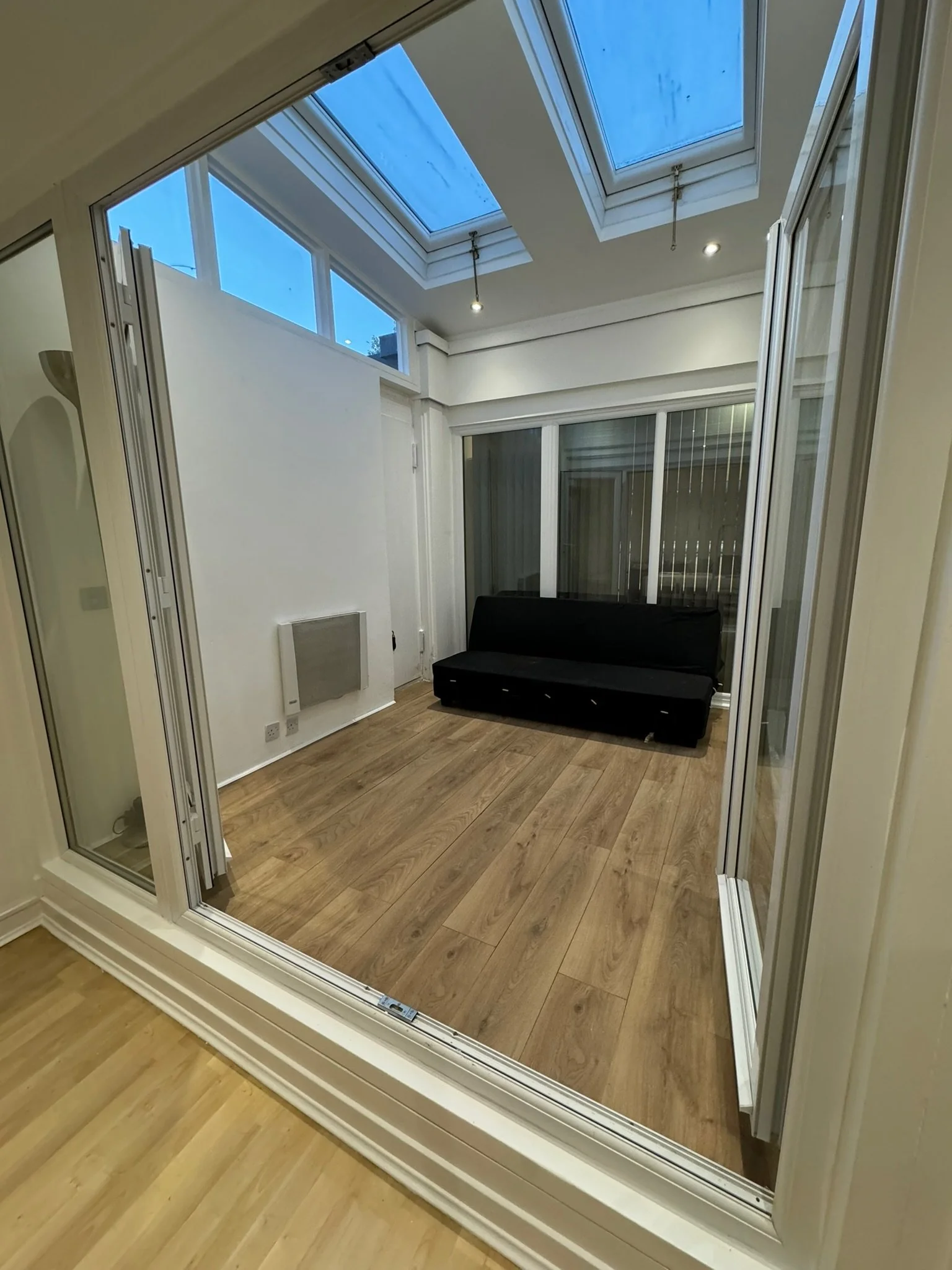 Empty room with wooden flooring, a black futon, large glass sliding doors, skylights, and a small electric wall heater.