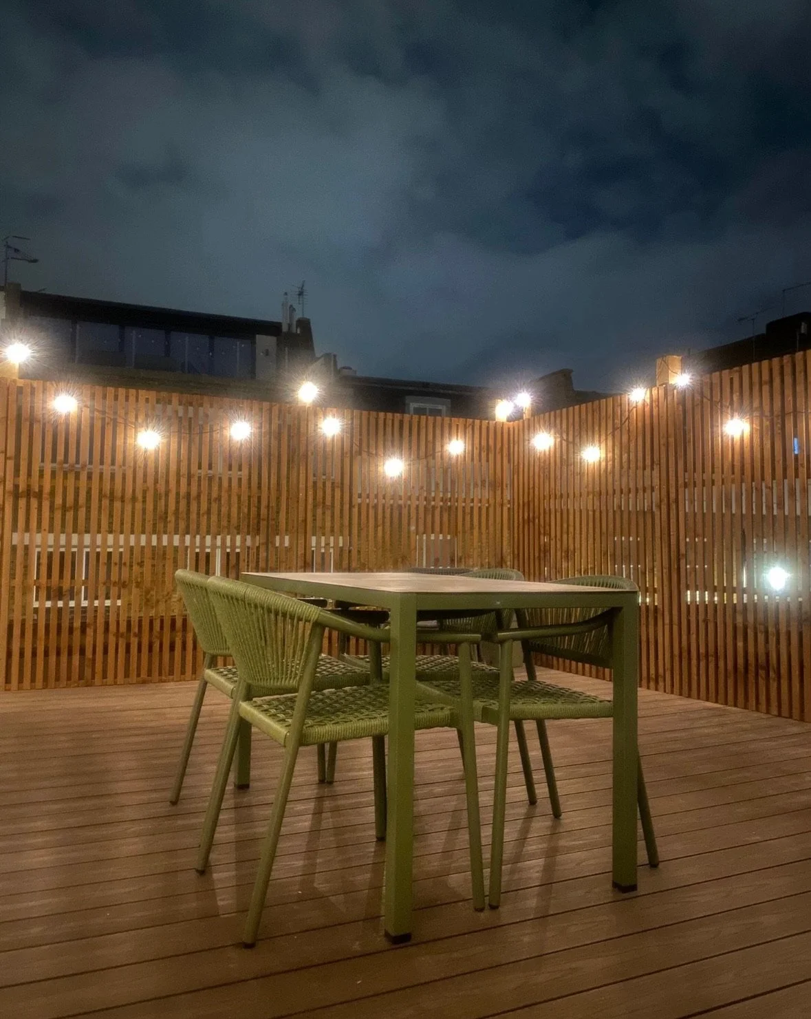 Empty outdoor patio with a wooden table and four green chairs, surrounded by a wooden privacy fence with string lights, under a dark cloudy sky at night.