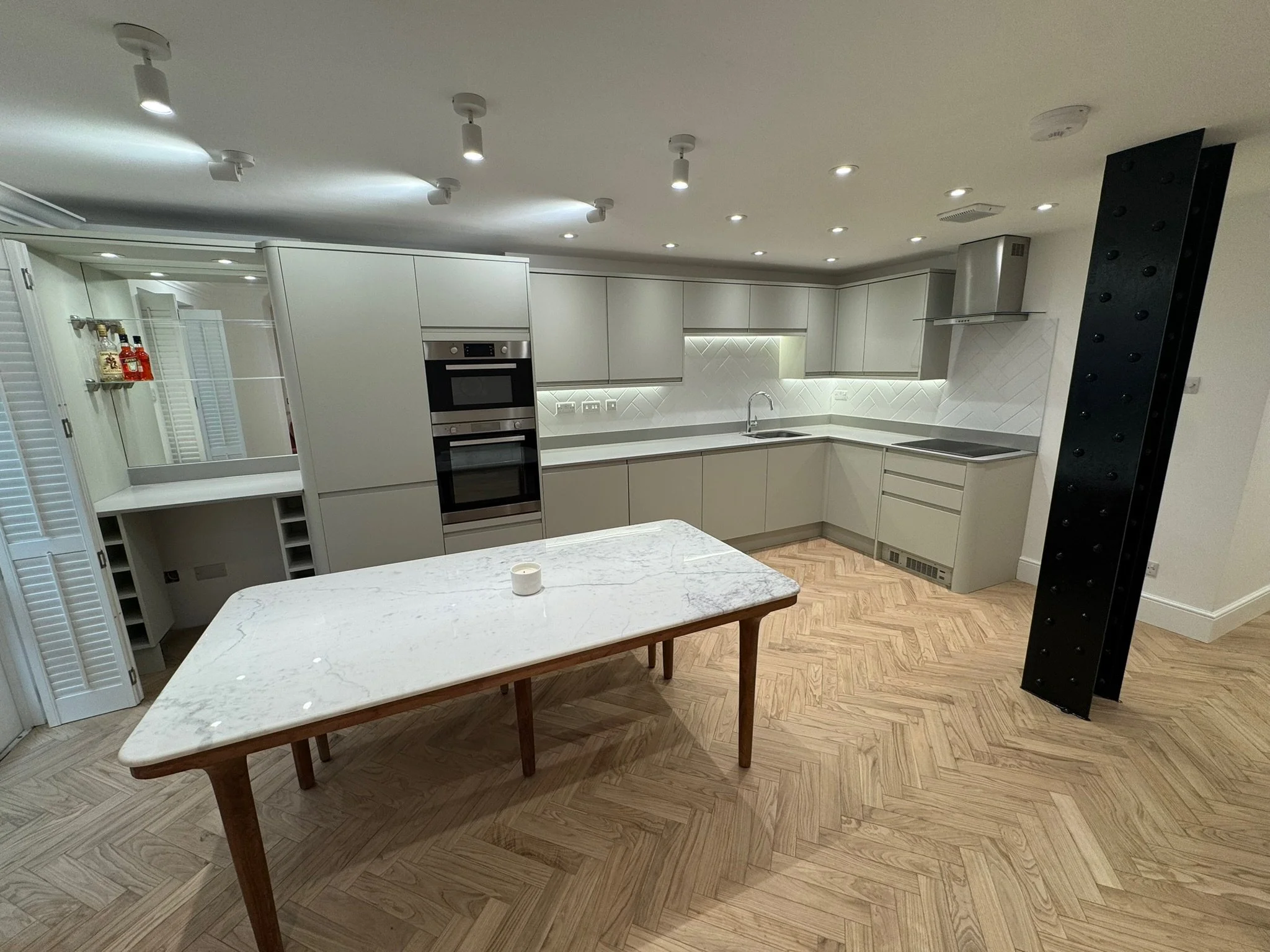 Modern kitchen with white cabinets, built-in dual ovens, marble-topped island, and wooden herringbone flooring.
