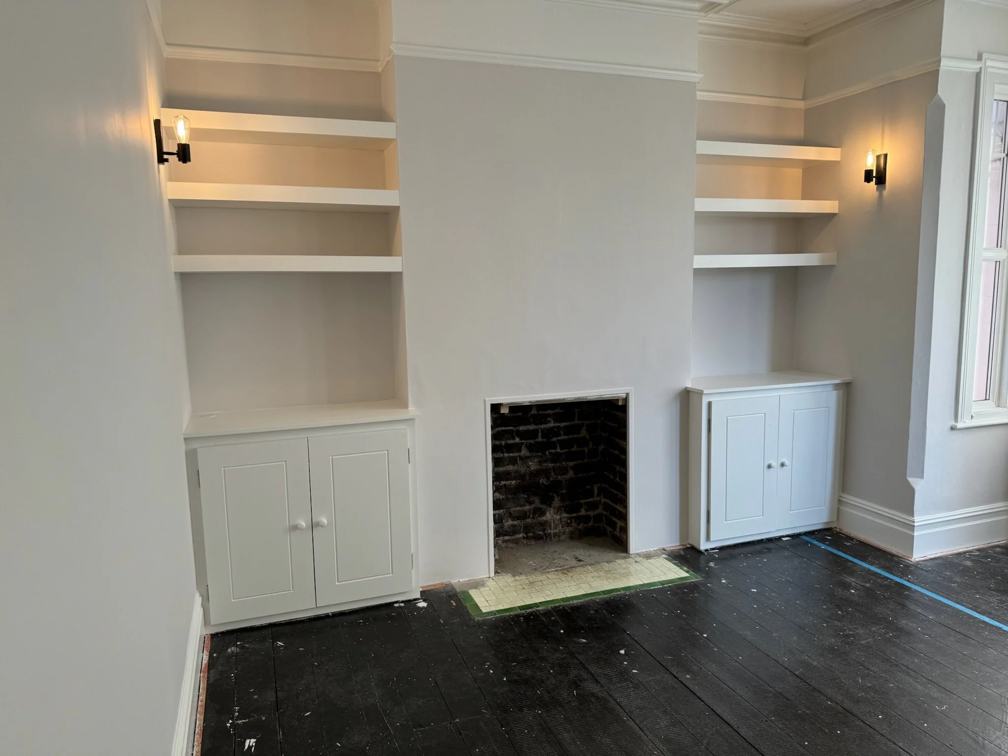 Living room with built-in white shelves and cabinets on either side of a fireplace opening, which is currently empty and partially under construction. There are two wall-mounted lights, dark wooden floors, and a window on the right side.