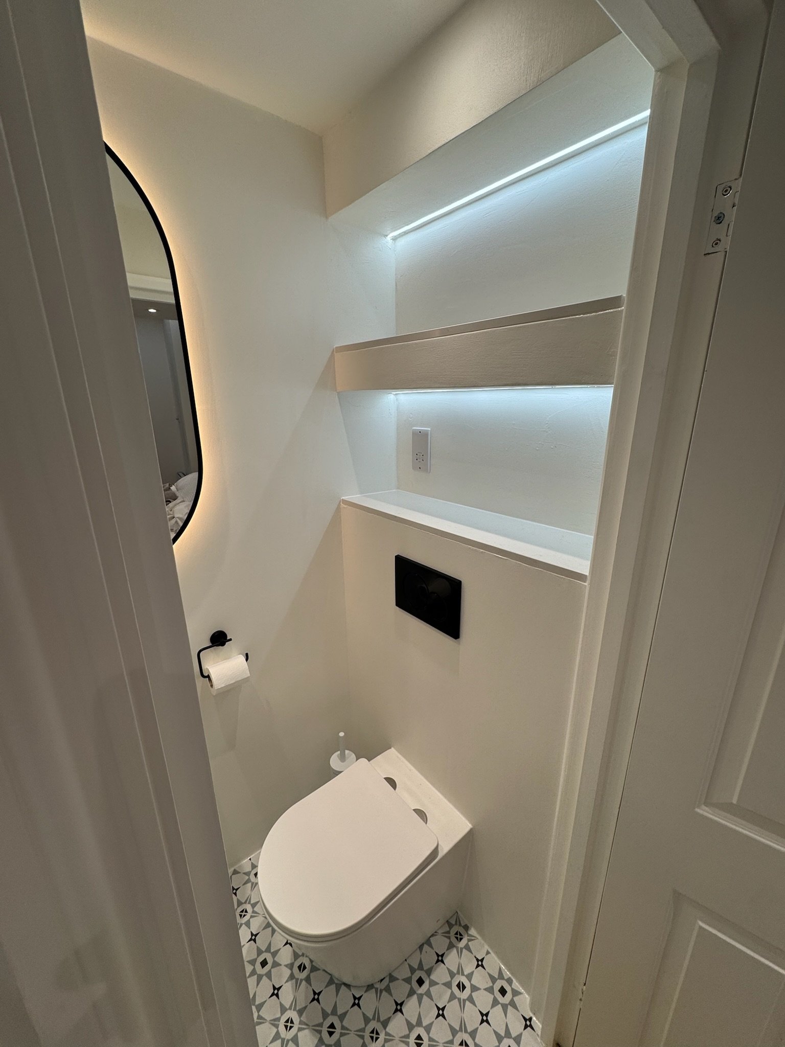 Modern bathroom with white walls, built-in shelves with lighting, a wall-mounted toilet, mirror, and patterned floor tiles.