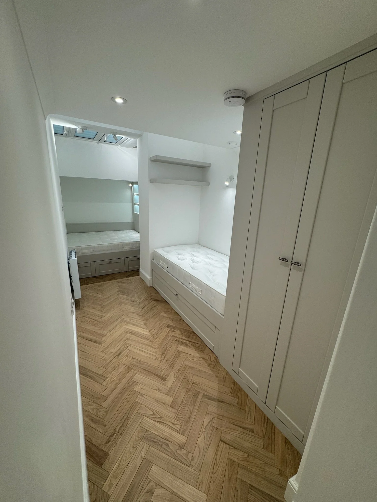 Small bedroom with white walls, wooden parquet floor, two shelves above a bed with white mattress, built-in wardrobe, skylight, and recessed ceiling lights.